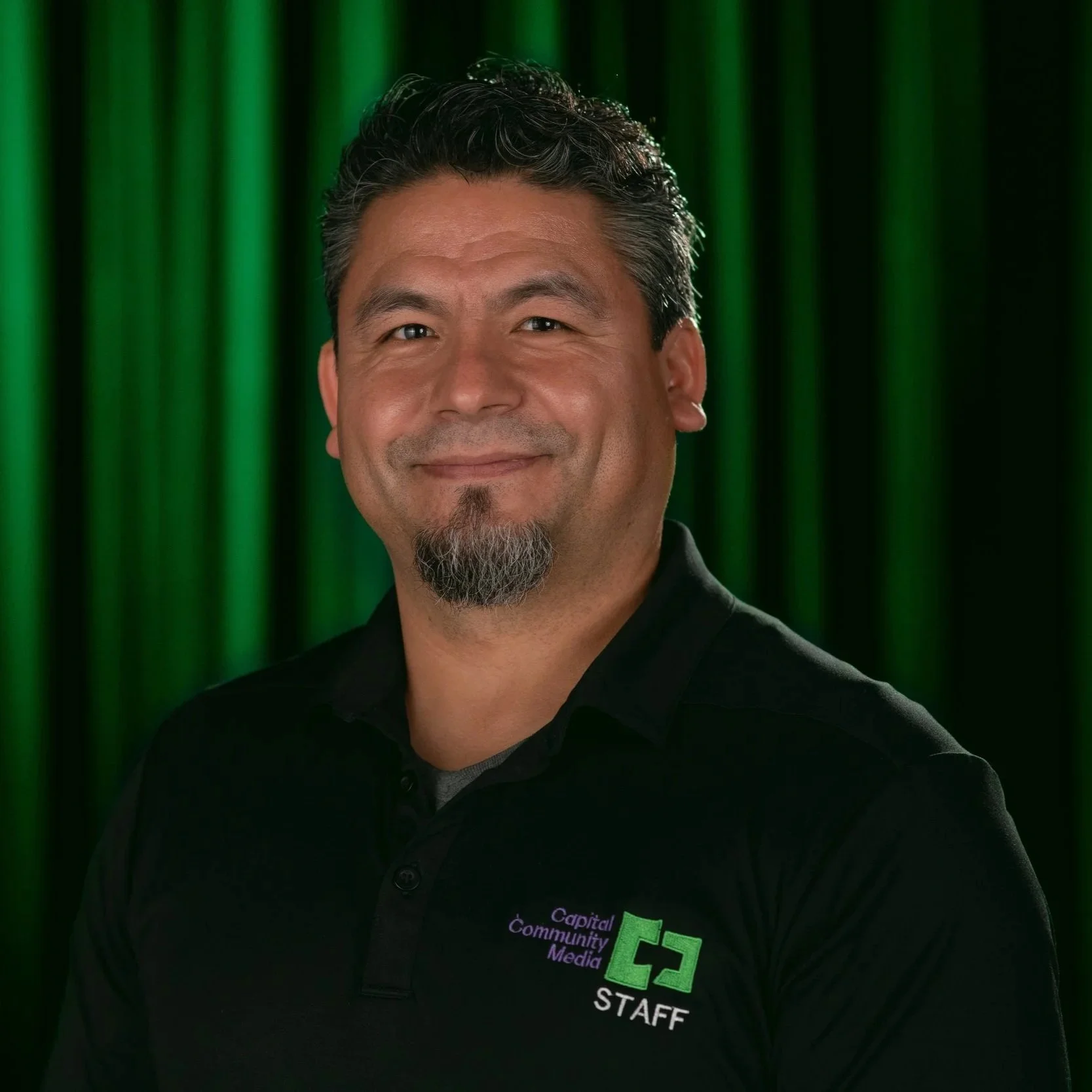 A man in black shit with Capital Community Media Staff on it with a back and Green background.