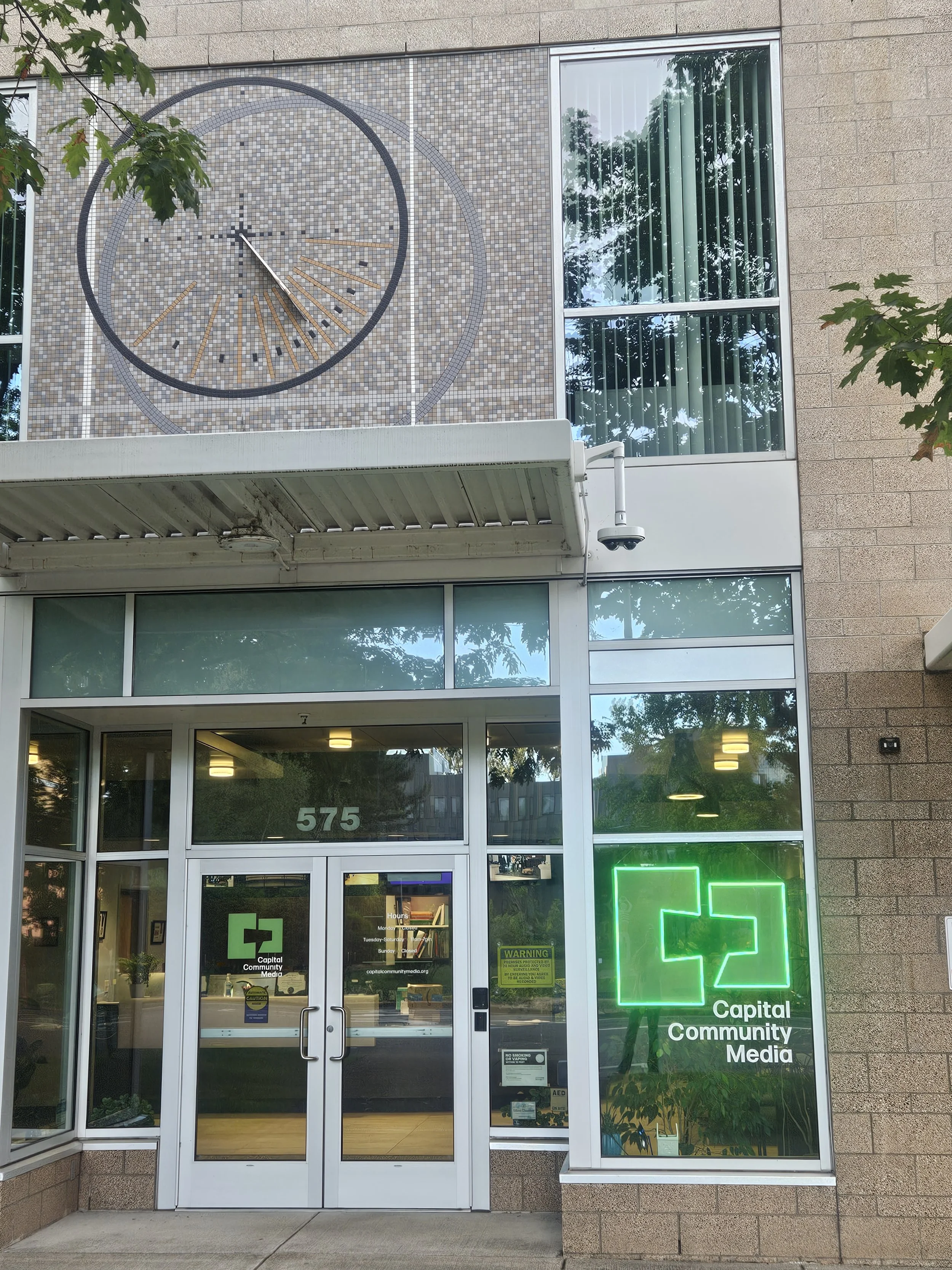 Picture of the front of Capital Community Media. Brick building with windows in the front and glass doors. Green Capital Community Media lighted logo to the right.