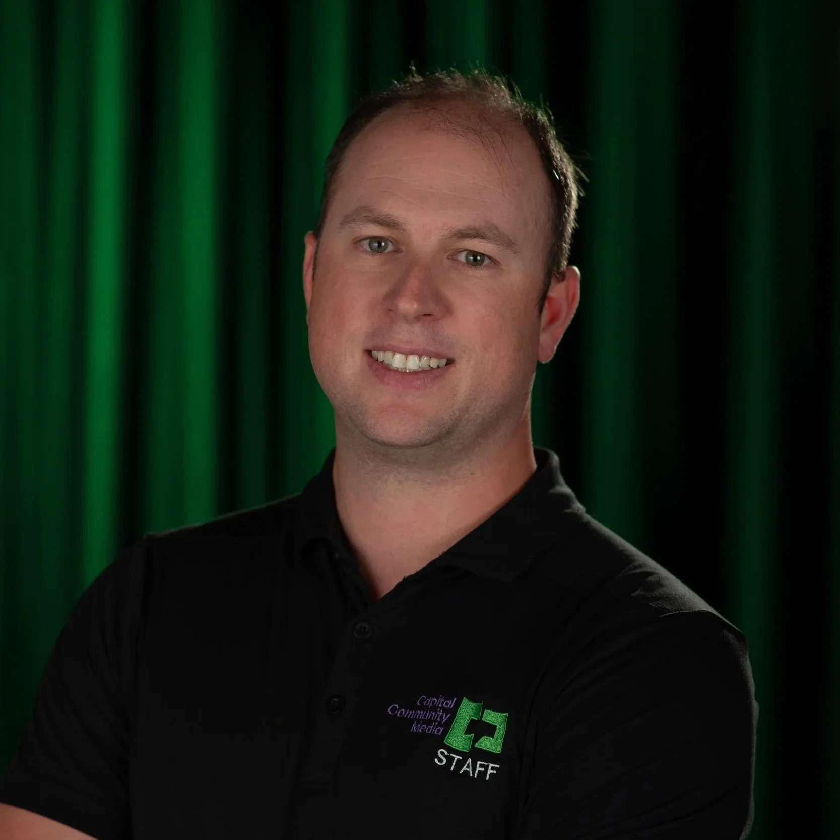 A Man in black shit with Capital Community Media Staff on it with a back and Green background.