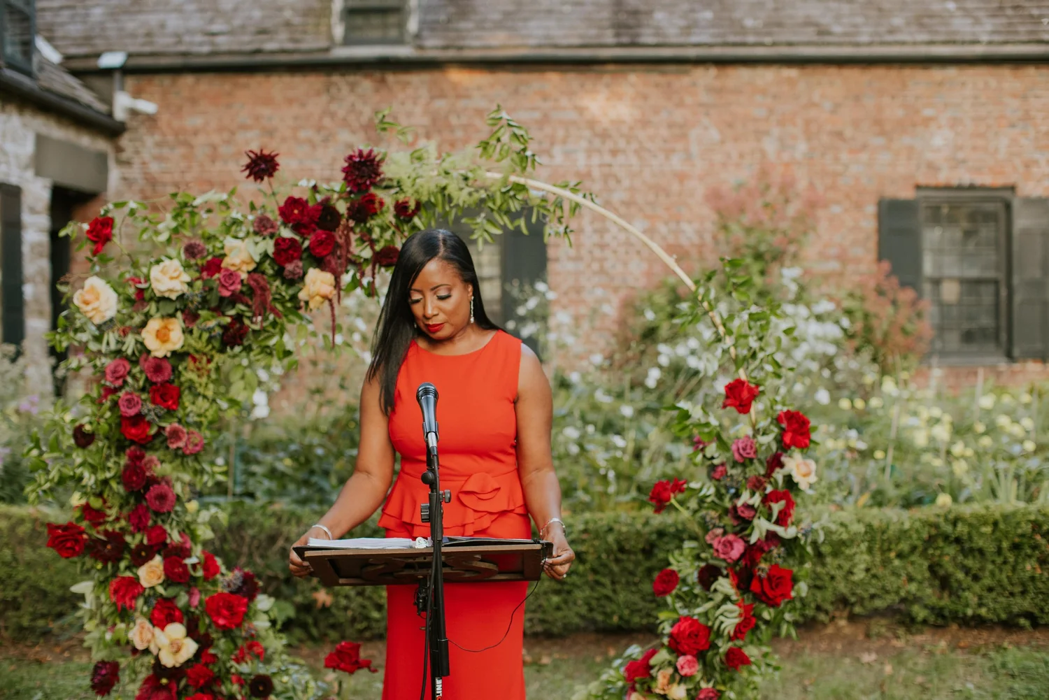 Aretha in a red gown reading at the lectern under a floral arch