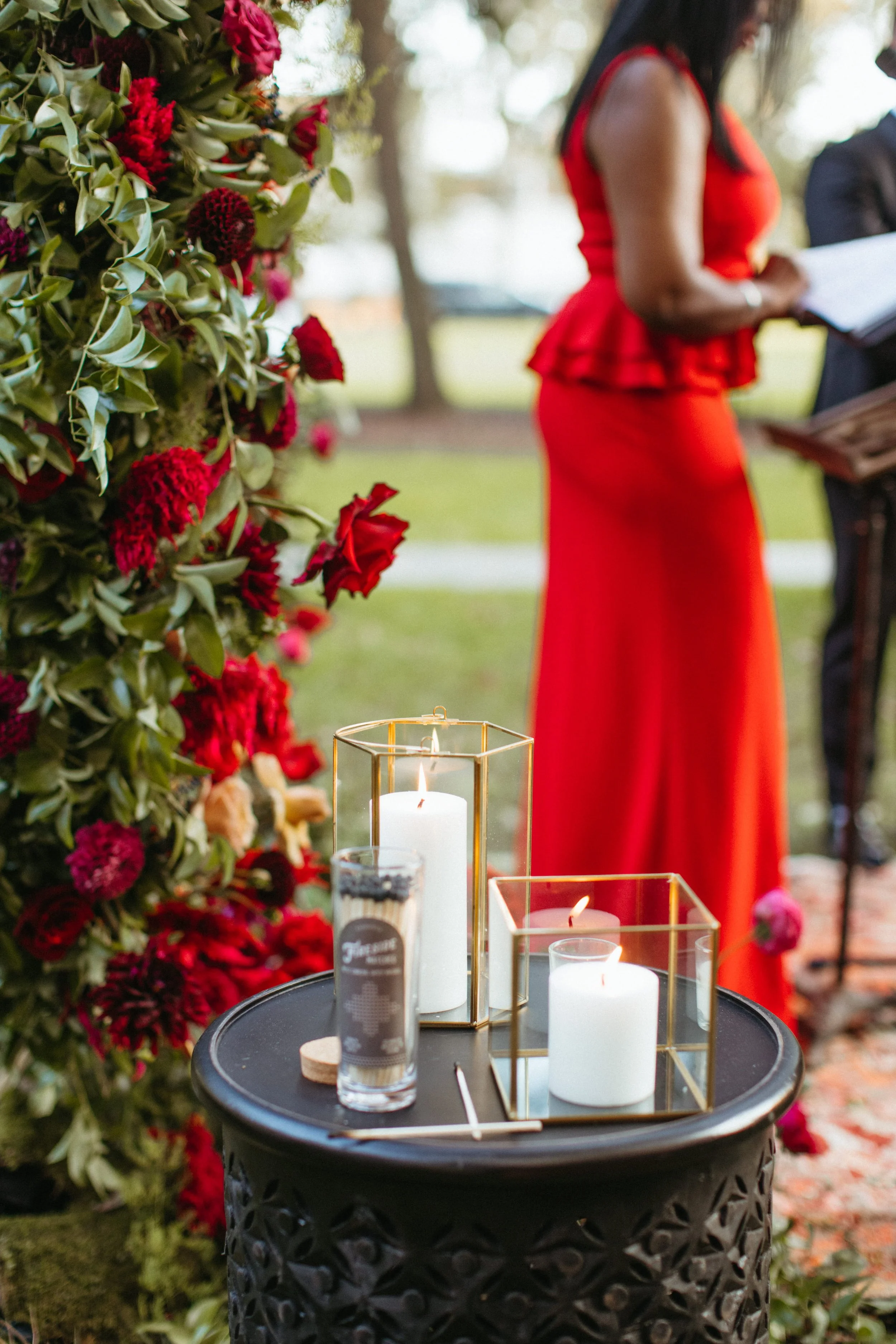 Candles in gold lanterns beside a red floral arch as officiant Aretha Gaskin, wearing a red dress, leads Matthew and Corey's wedding ceremony at Senate Garage in Kingston, New York