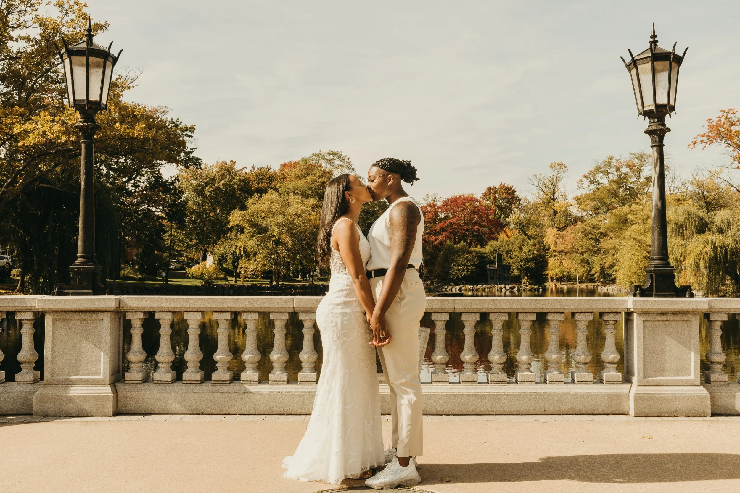 Jaide and Amiaya kiss while holding hands beside the pond during their fall wedding at Mindowaskin Park in Westfield, NJ.