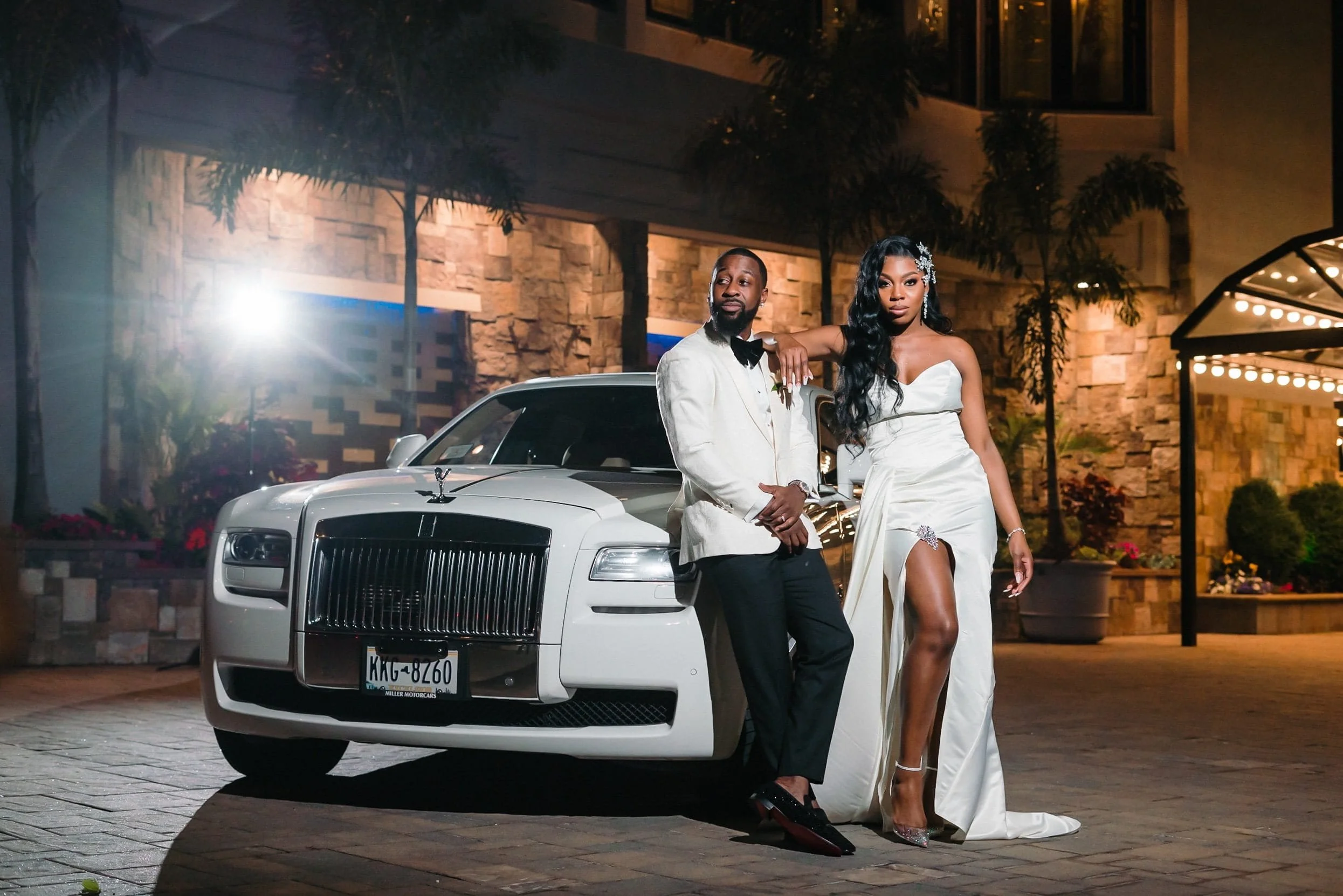 Bride in a strapless gown and groom in a white tuxedo jacket posing beside a white Rolls-Royce outside The Sand Castle in Franklin Square, NY