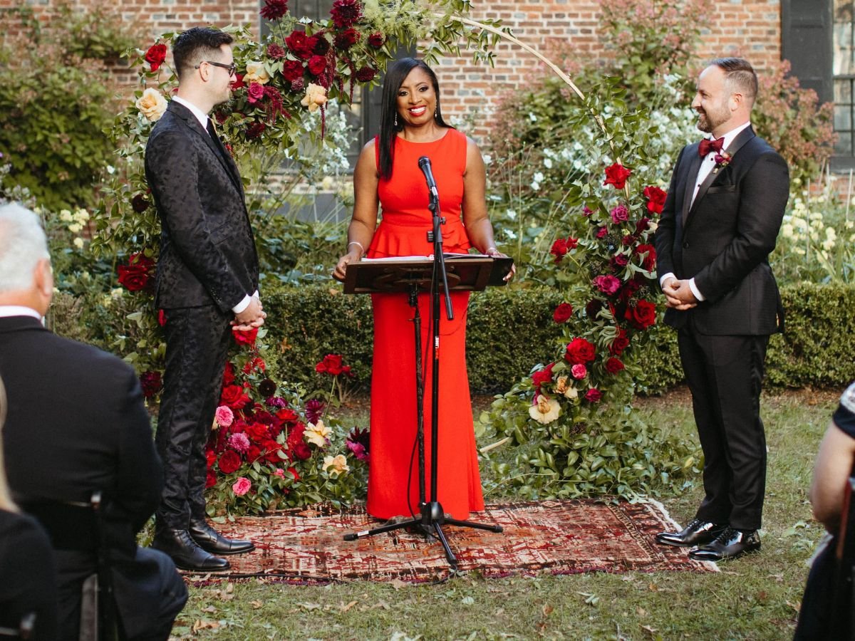 Wedding officiant in a red dress coordinated to the couple's colors during their ceremony at Senate House Grounds in Kingston, New York