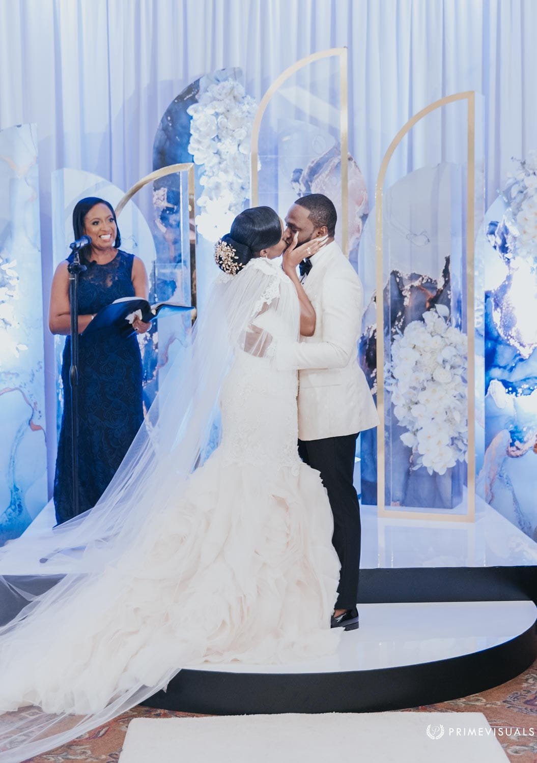 Officiant Aretha Gaskin smiles at the microphone as the couple shares their first kiss in front of a white and blue floral backdrop