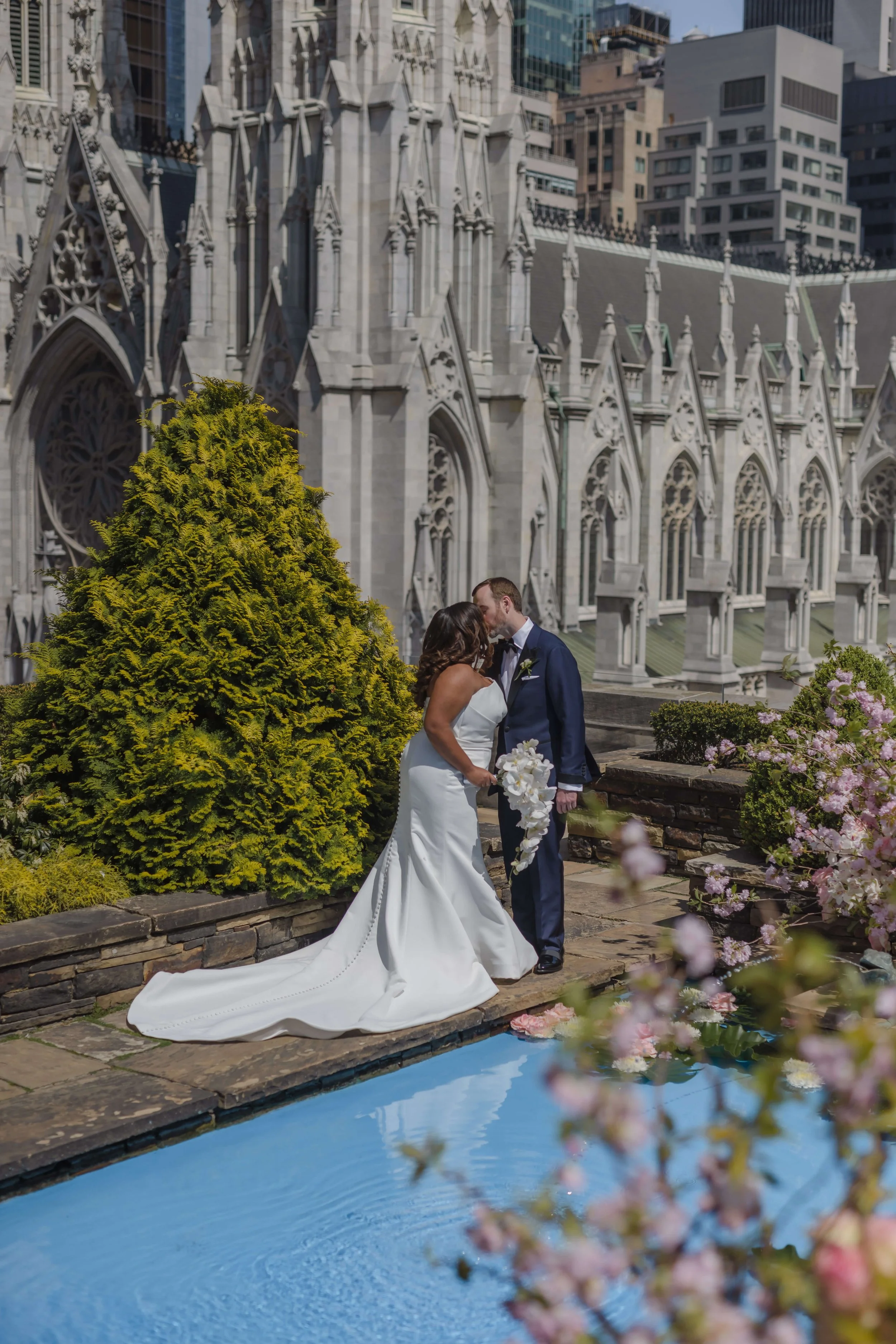 Justine and Steve share a kiss beside a reflecting pool in a rooftop garden with a Gothic cathedral rising behind them