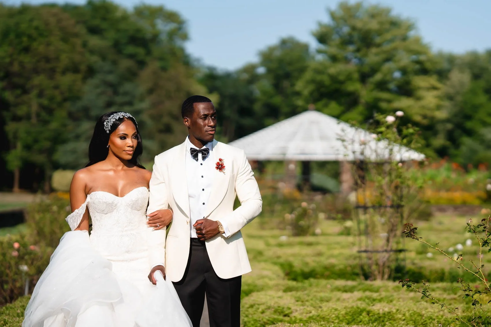 Bride in an off-the-shoulder gown walking arm in arm with her groom in a white tuxedo jacket at Deep Cut Gardens in Middletown, NJ