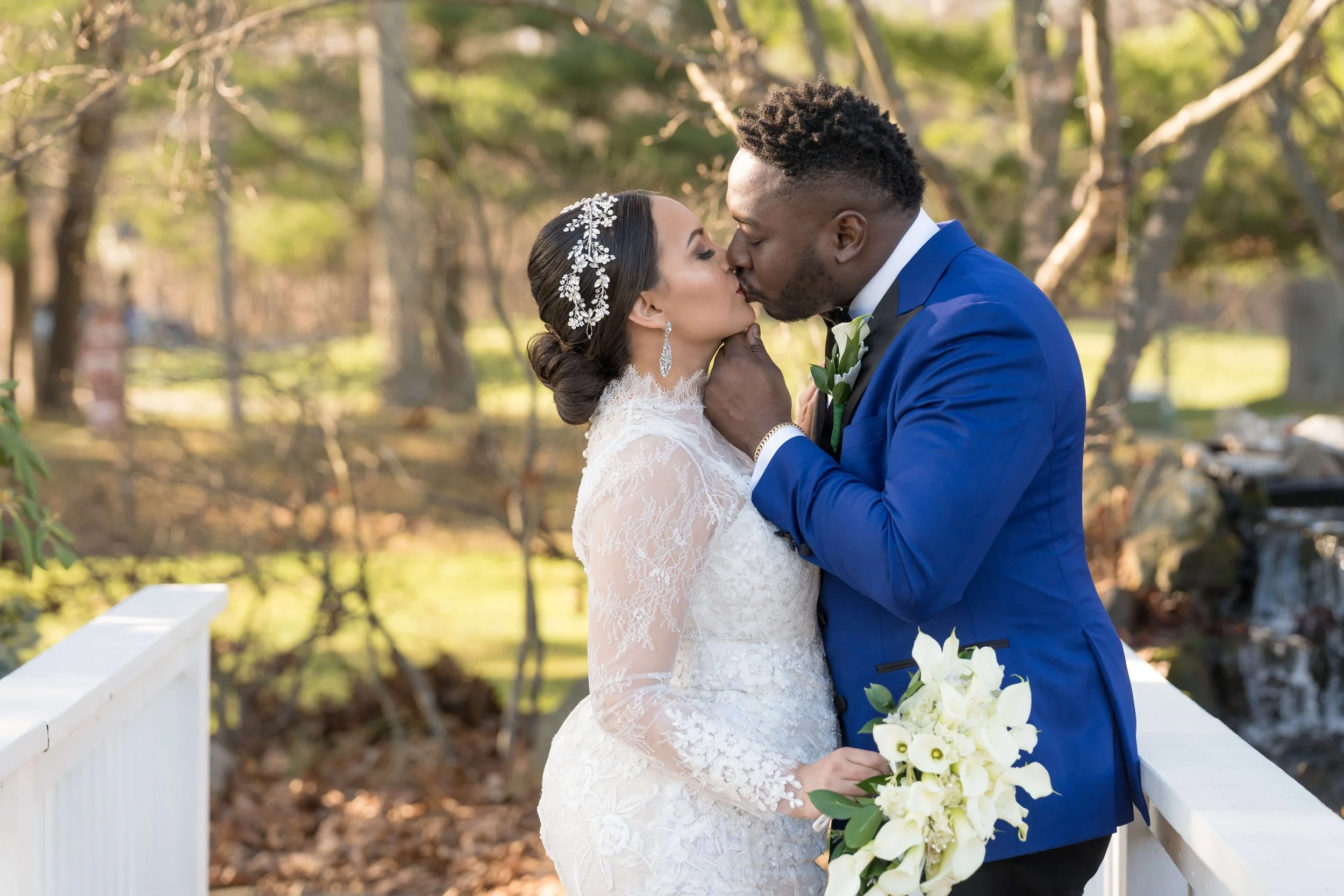 Groom in a royal blue tuxedo jacket kissing his bride in a long-sleeve lace gown on a white footbridge at Stonebridge Country Club in Smithtown, NY