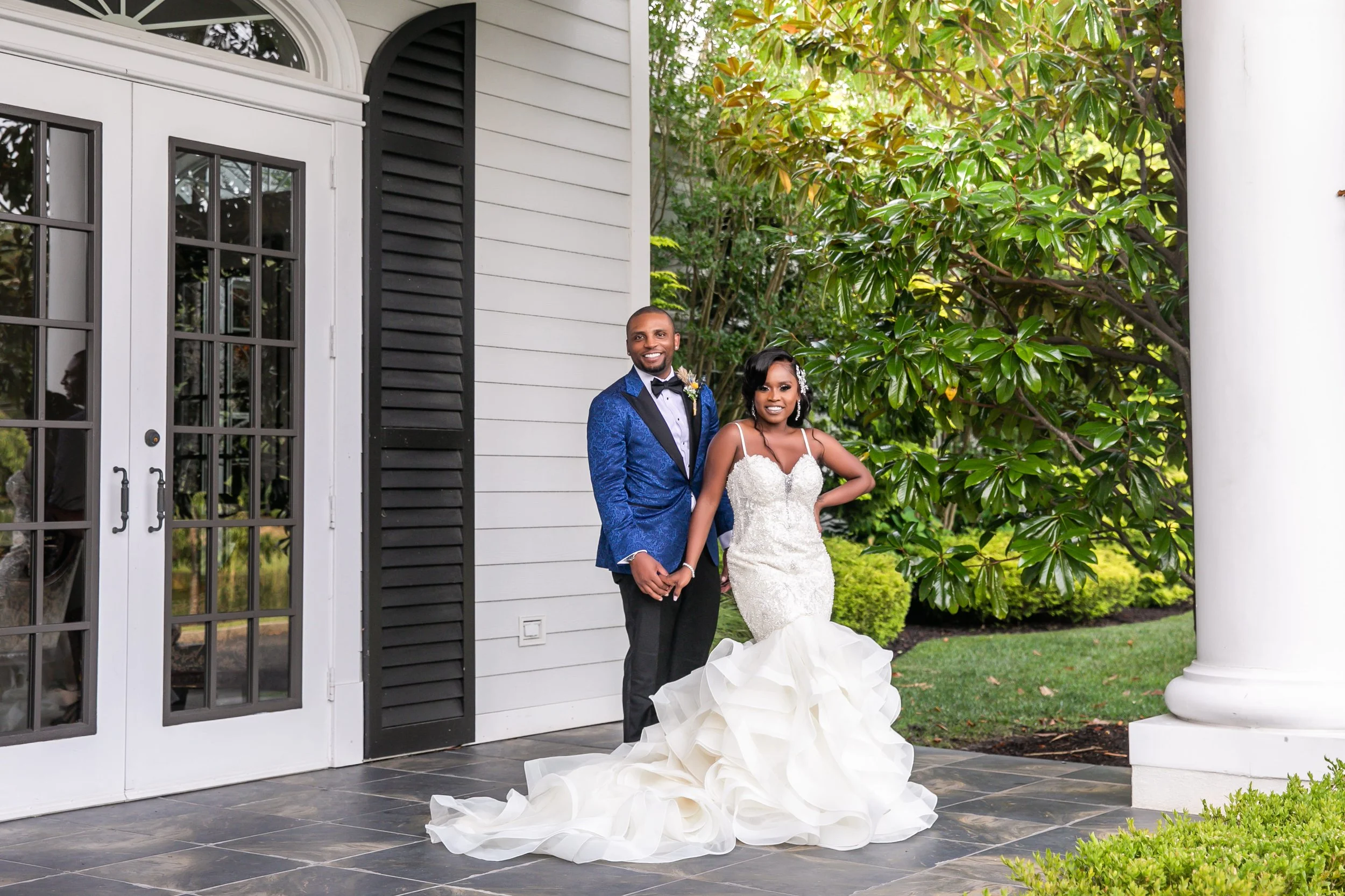 Bride in a beaded mermaid gown with a ruffled train holding hands with her groom in a royal blue tuxedo jacket on the porch at The Carriage House in Galloway, NJ
