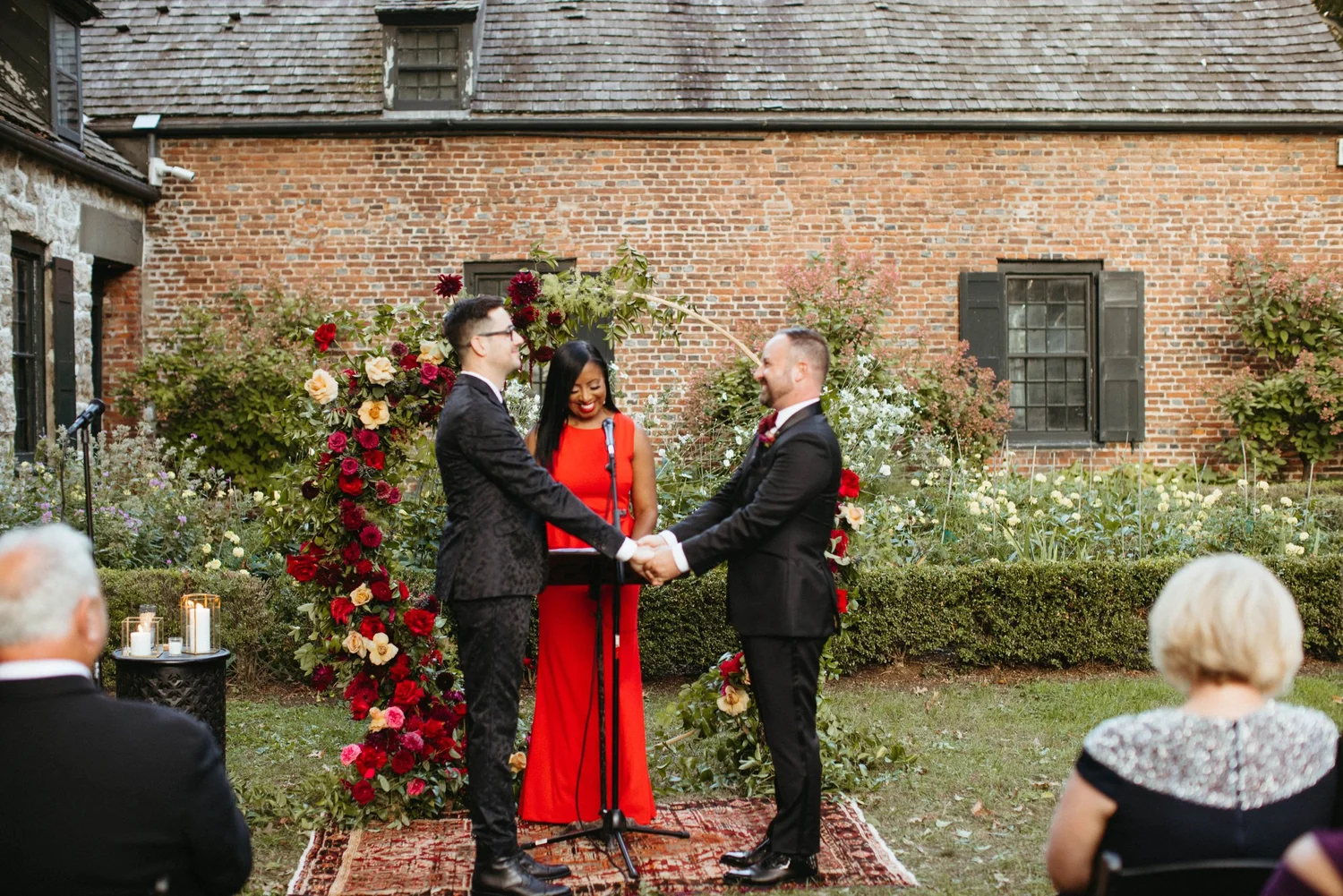 Aretha in a red gown officiating a garden wedding ceremony for two grooms