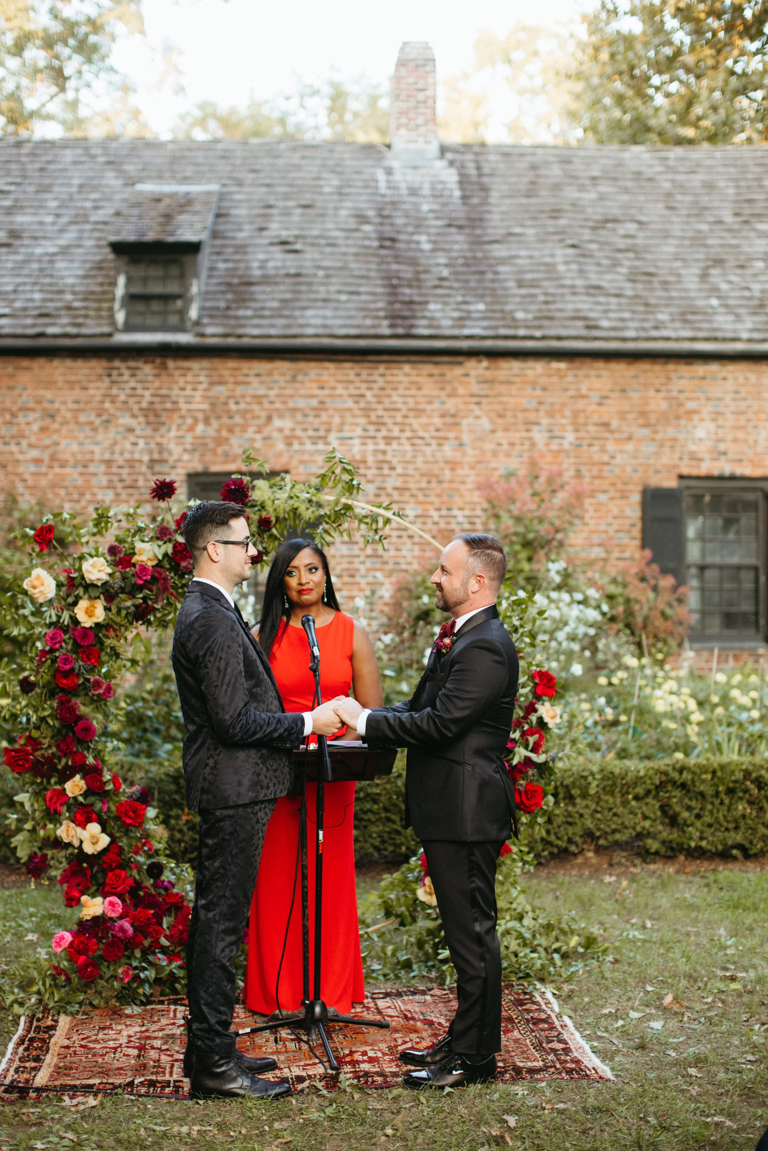 Wedding officiant in a red dress embracing a couple after their ceremony at Senate House Grounds in Kingston, New York