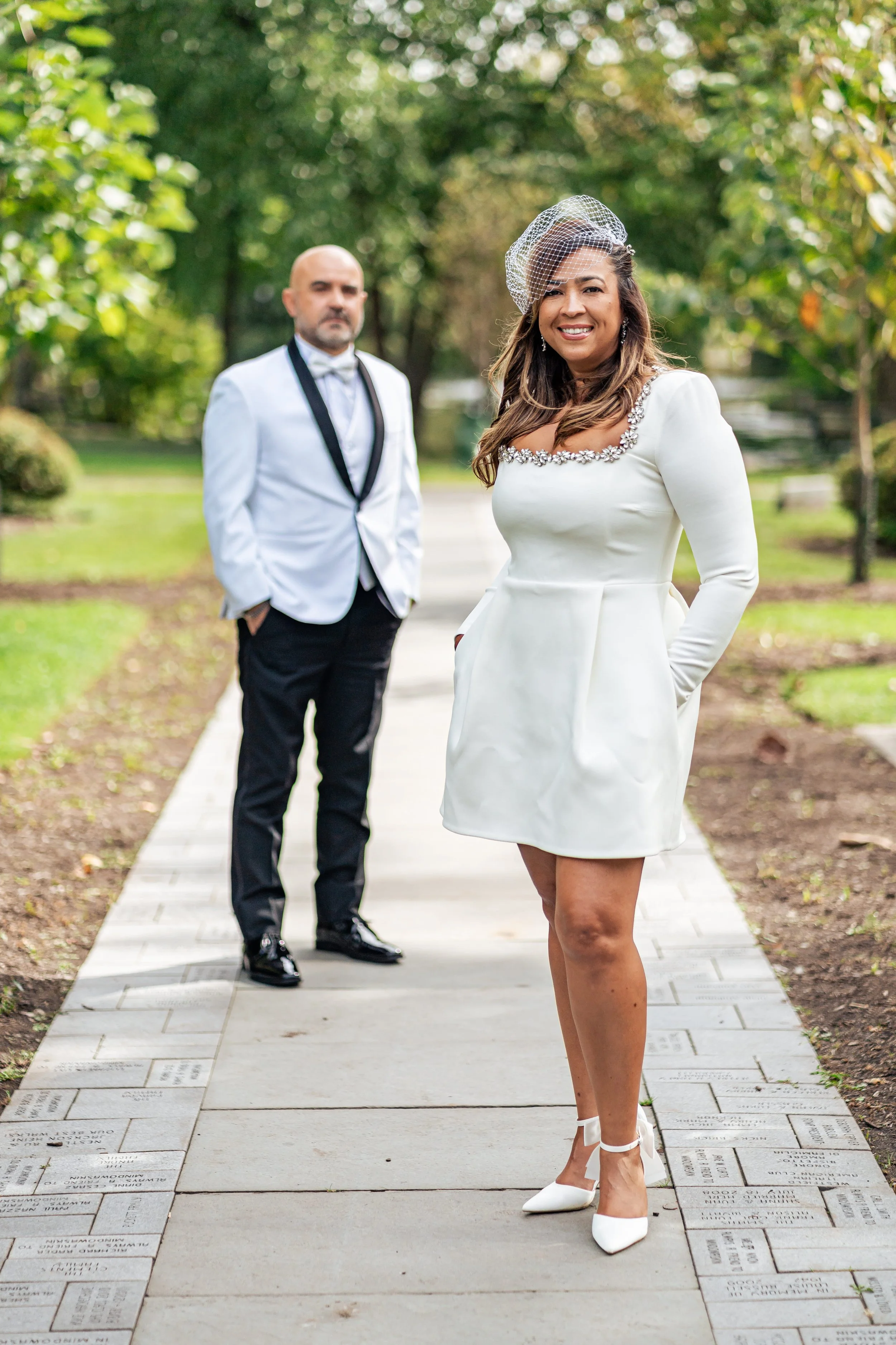 Lissette smiles in a short white dress and birdcage veil on a park path as Jack waits behind her at Mindowaskin Park in Westfield, NJ