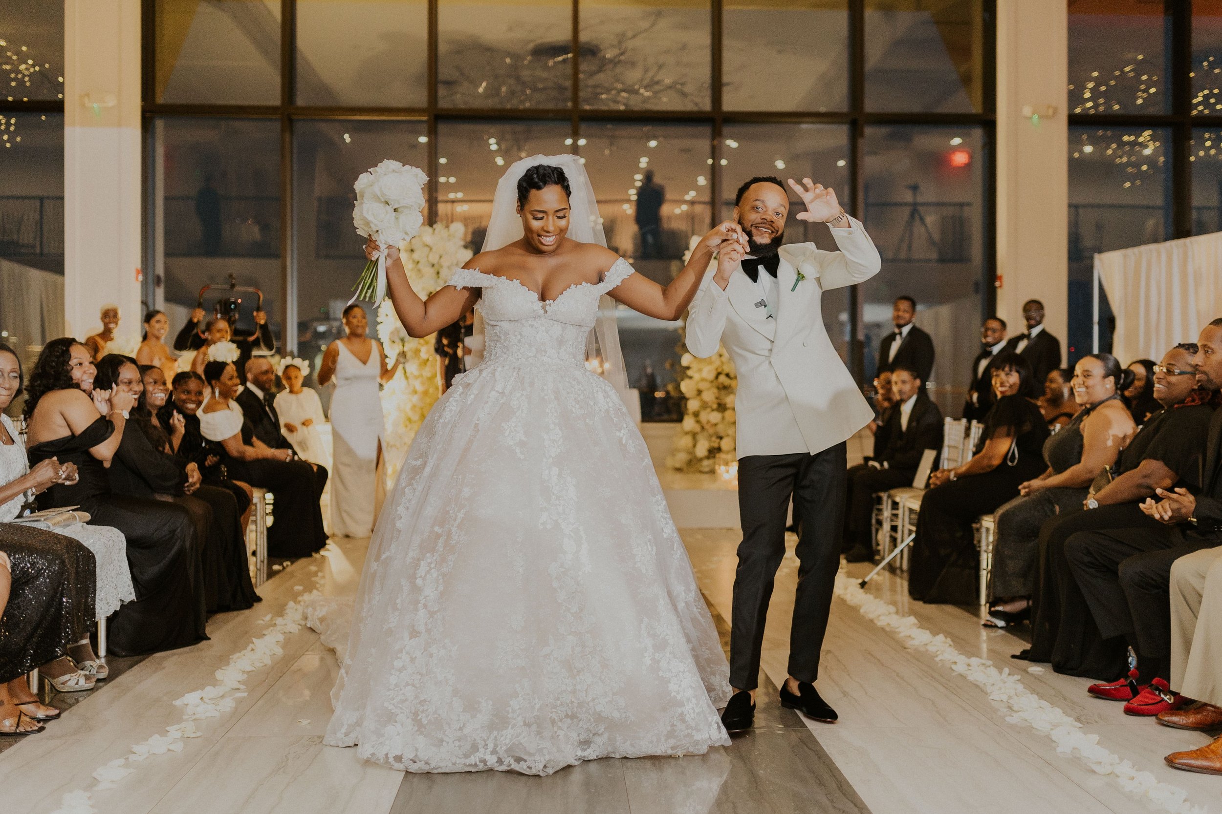 Bride raising her white rose bouquet as she and her groom in a white tuxedo jacket celebrate down the aisle after their ceremony at Hudson House in Jersey City, NJ