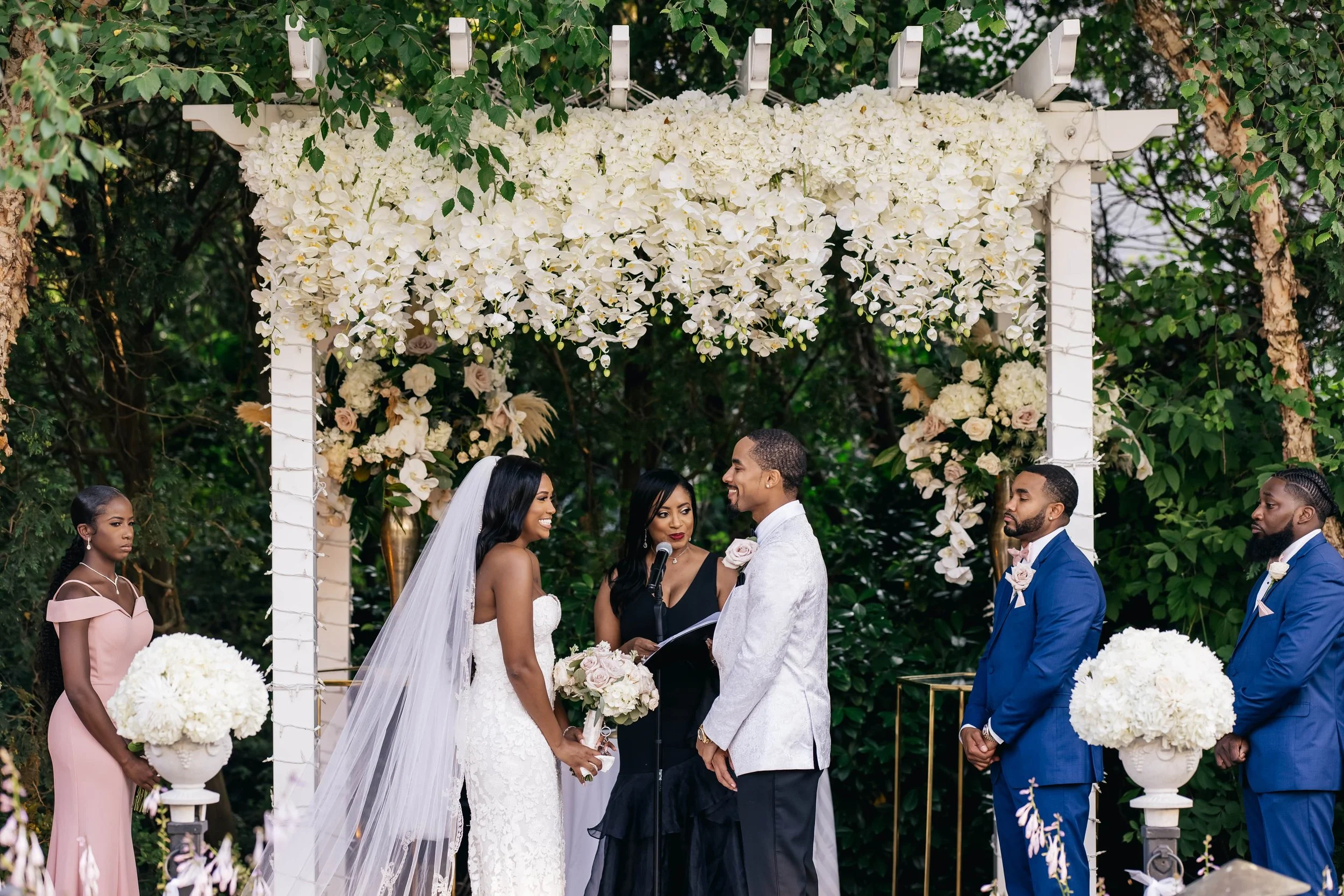 Wedding officiant standing with a couple beneath a floral arch during their ceremony at The Fox Hollow in Woodbury, New York