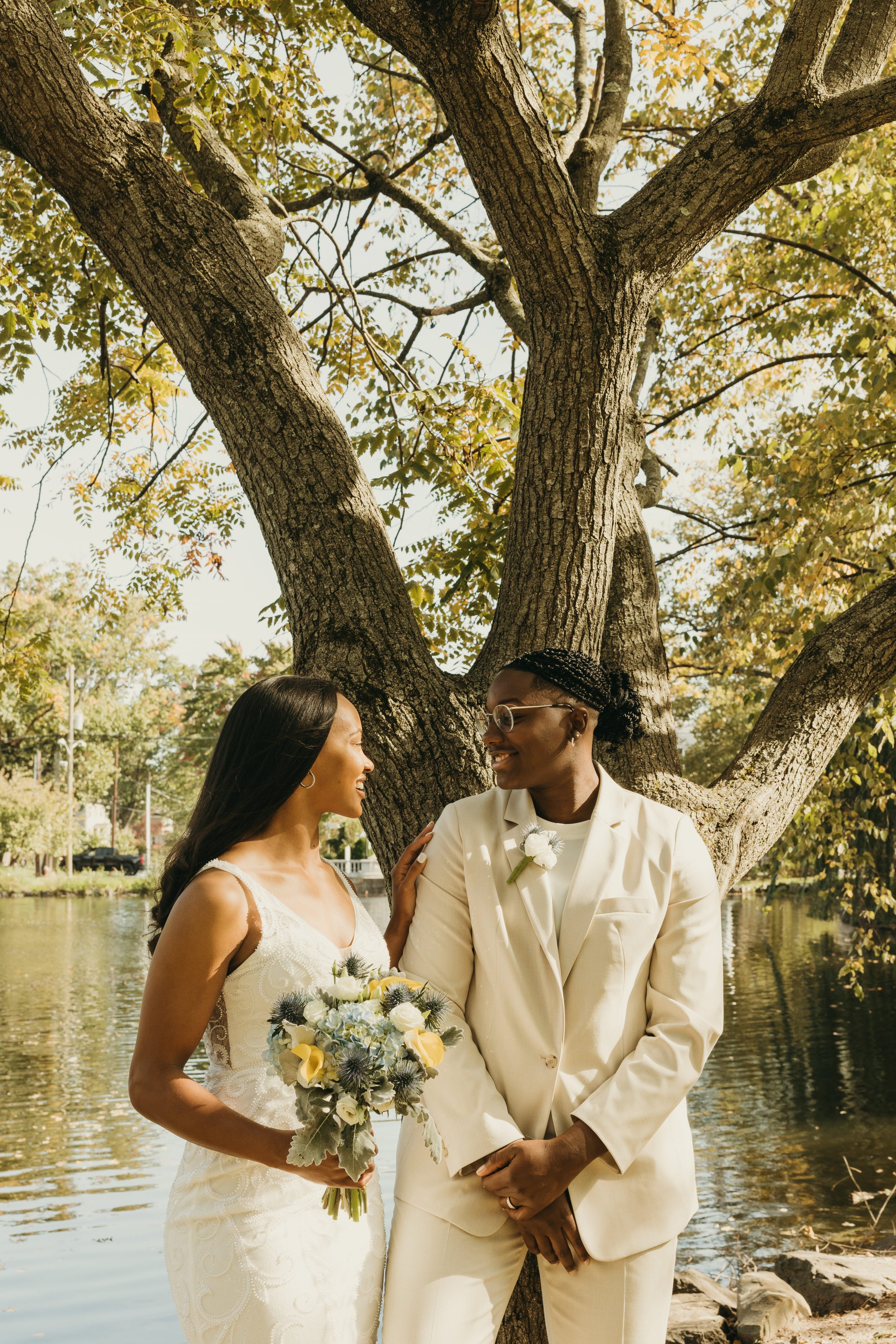 Jaide and Amiaya smile at each other beneath a large tree beside the pond during their fall wedding at Mindowaskin Park in Westfield, NJ.