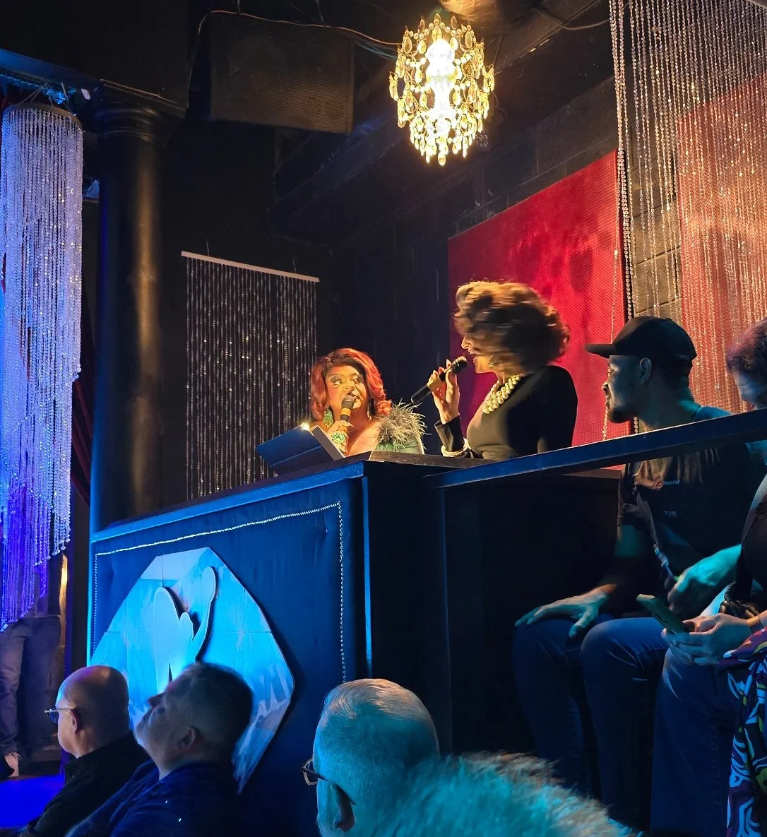  Two drag performers, former titleholders Kiara Mel (left) and Emory Starr (right) emcee the talent competition. They are sitting behind an emcee booth elevated off the side of the stage, speaking into microphones, with an audience watching. Sparklin