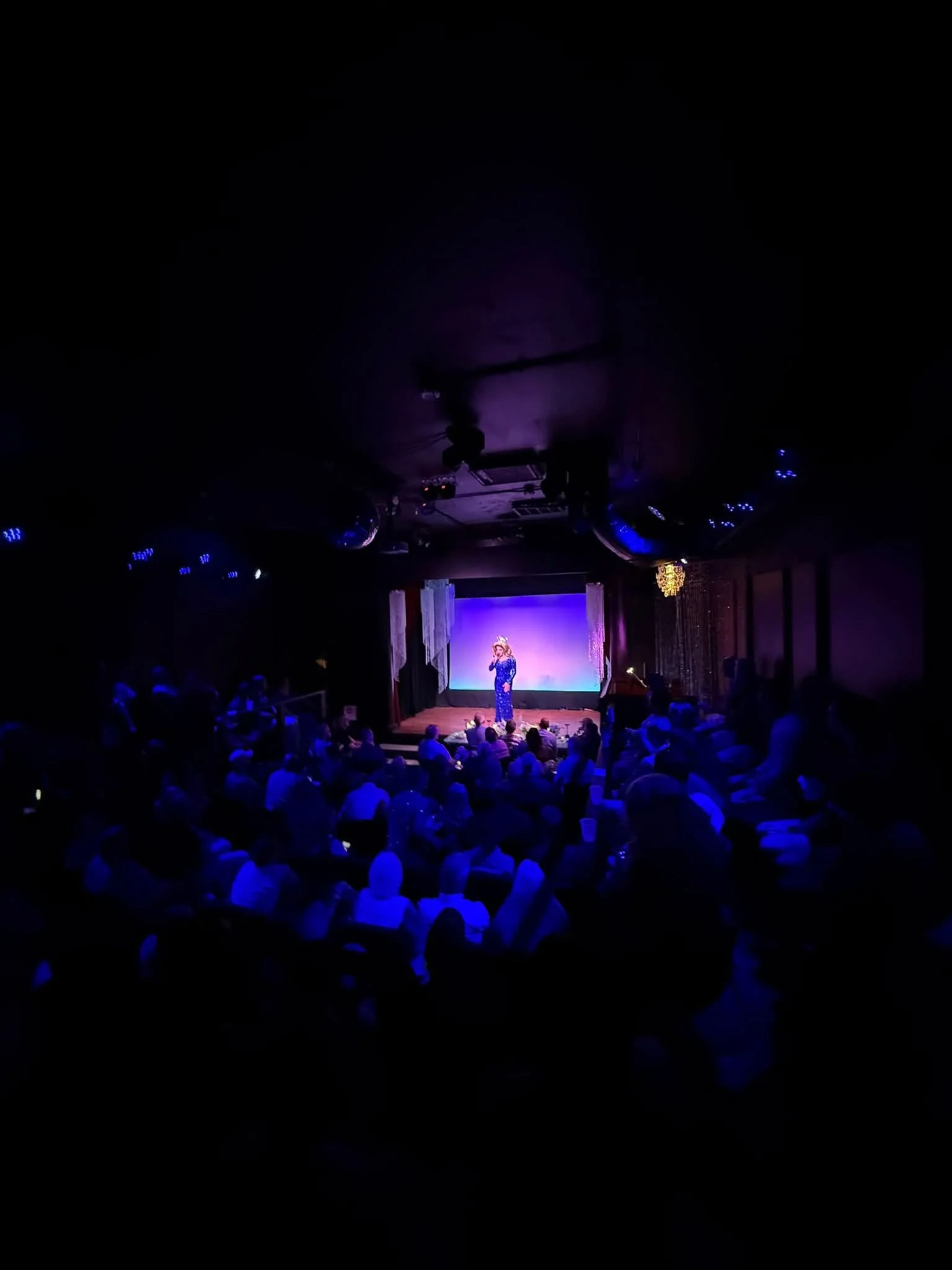  Tracy La Louisianne, Miss Gay America 2026, stands alone on a brightly lit stage, facing a large seated audience in a dark theater. Blue lighting illuminates the crystal columns flanking the stage, creating a dramatic atmosphere. 