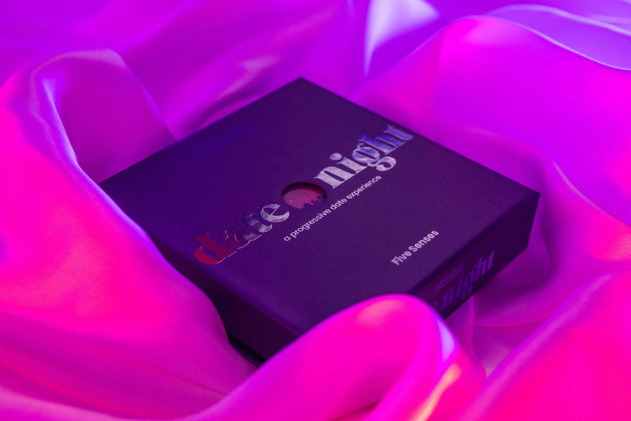 Blue box with the words "Date Night a progressive date experience" lays on a silk sheet. The lighting is moody and colored.