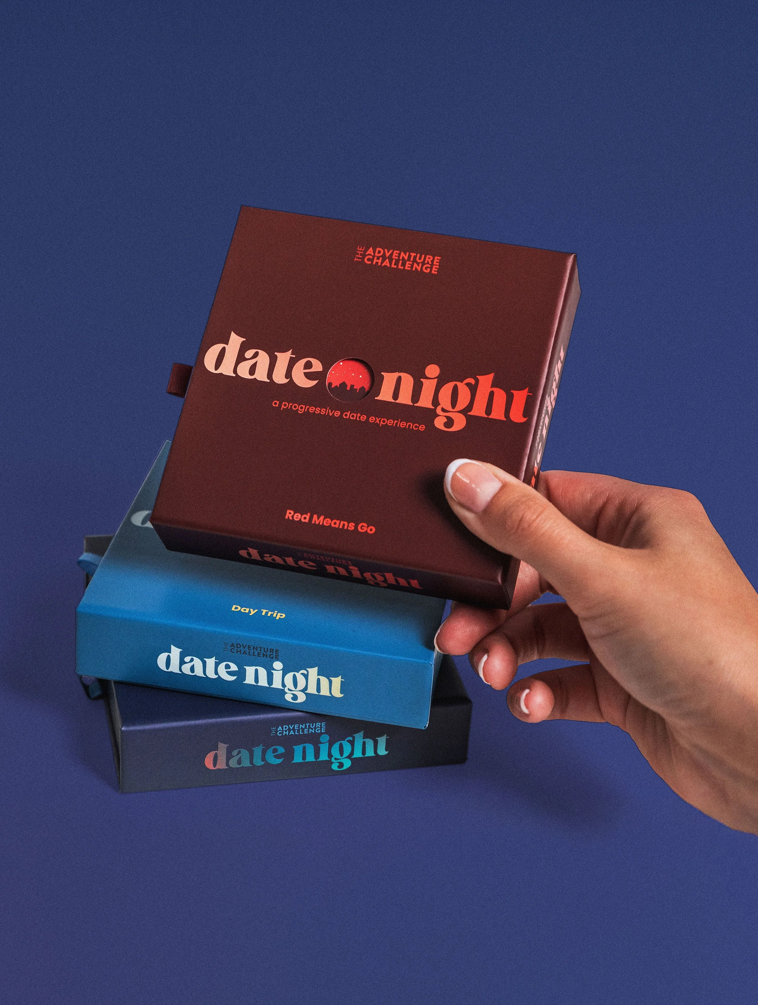 Three product boxes sit against a blue backdrop. A hand holds one of the boxes that is a red box. The other two boxes are light blue and indigo blue. Each box has ombre colored words that say "Date Night a progressive date experience."
