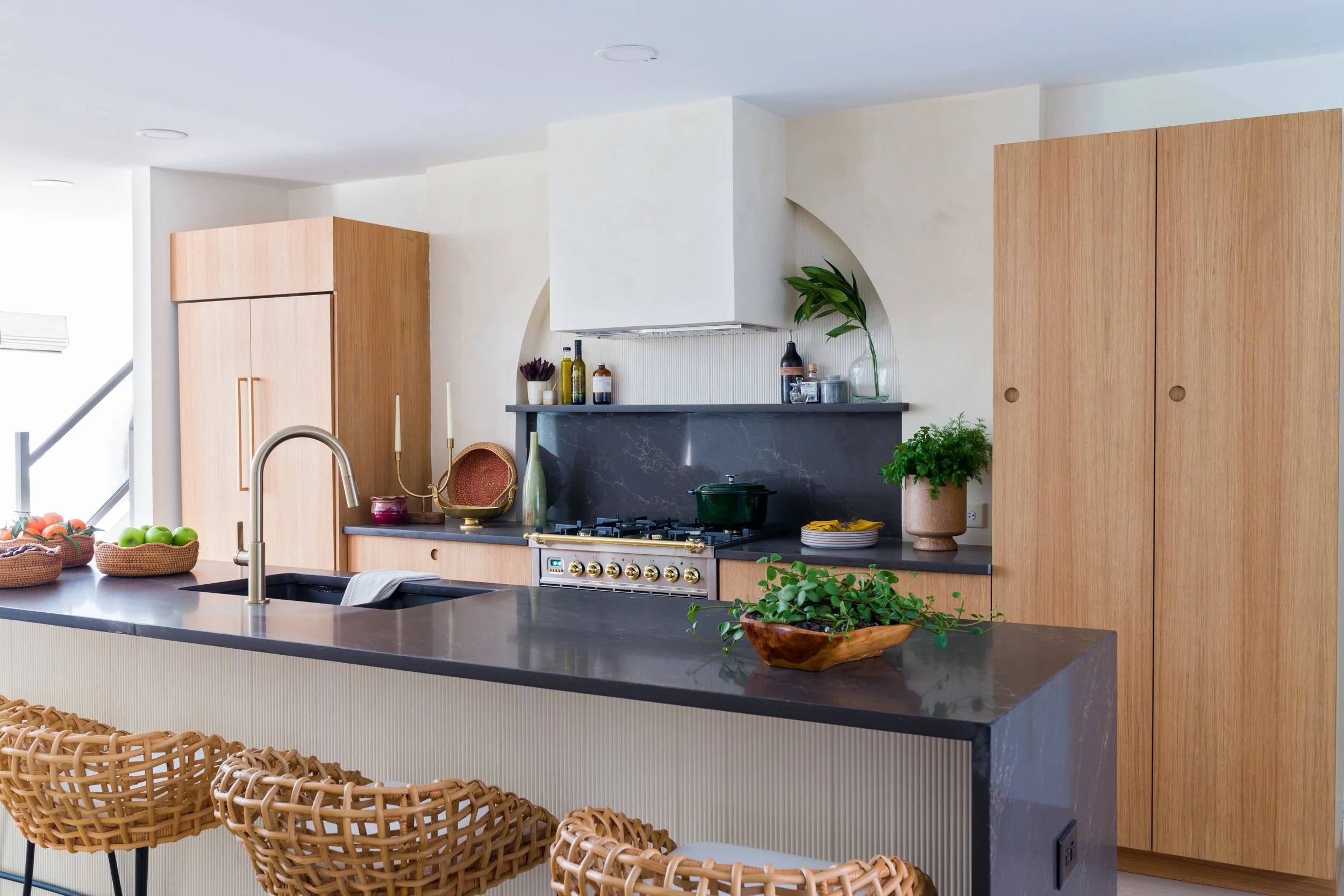 Modern kitchen with fluted island, light wood cabinetry, arched range surround, and dark stone countertops.