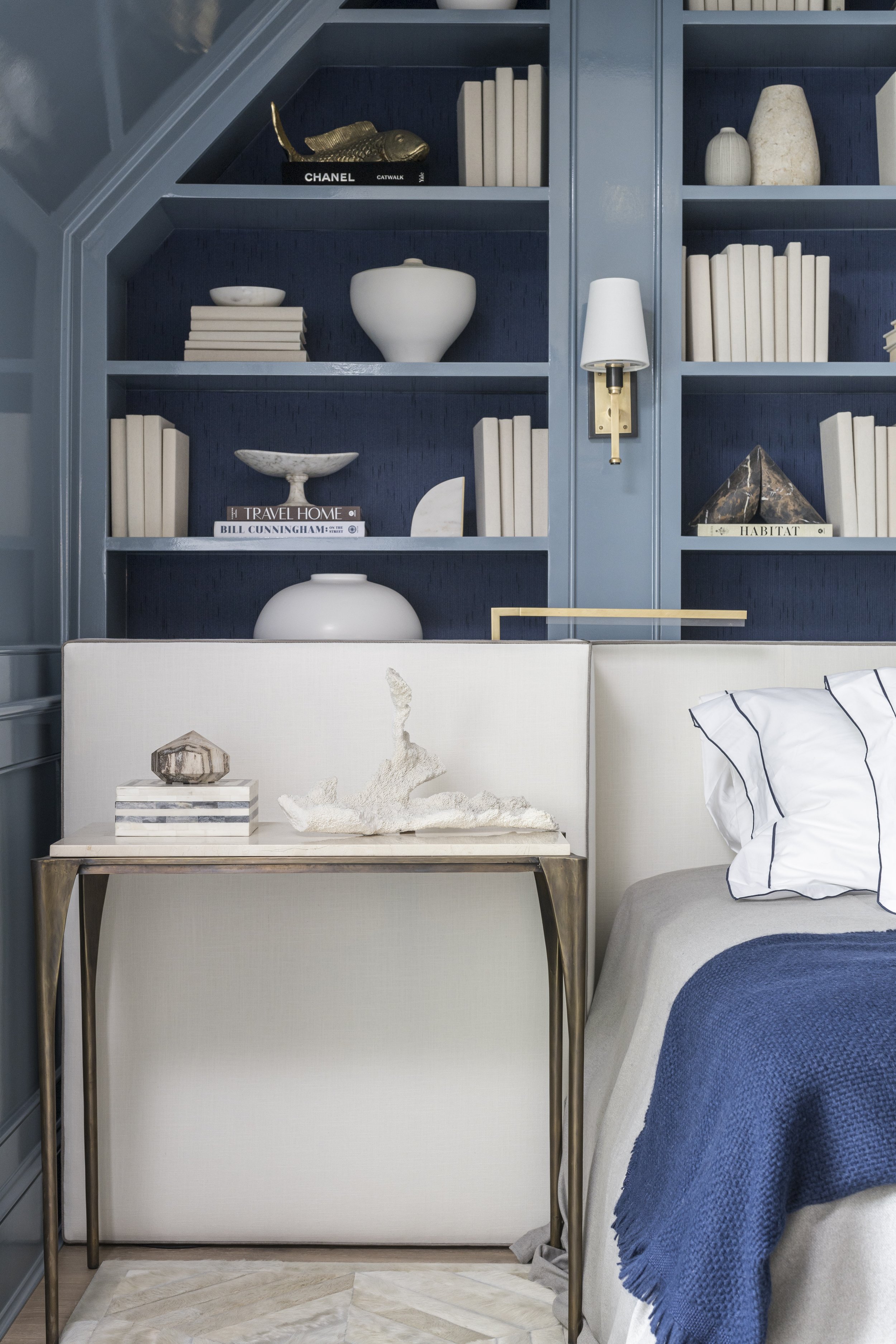 Bedroom detail with built-in blue bookcase, upholstered bed, brass wall sconce, and styled night table.