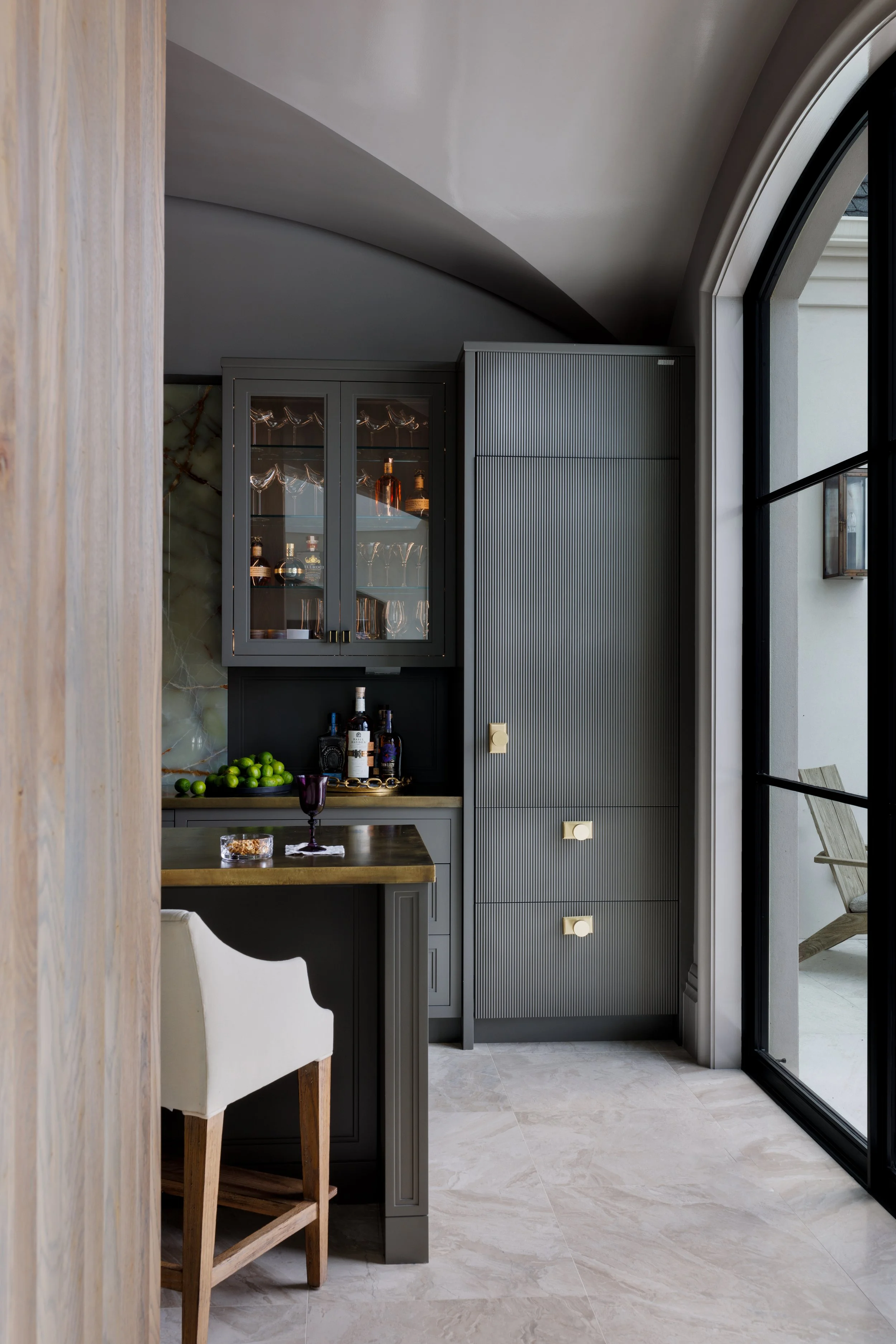 Custom kitchen bar with fluted cabinetry, glass-front cabinets, stone countertop, and arched doorway.