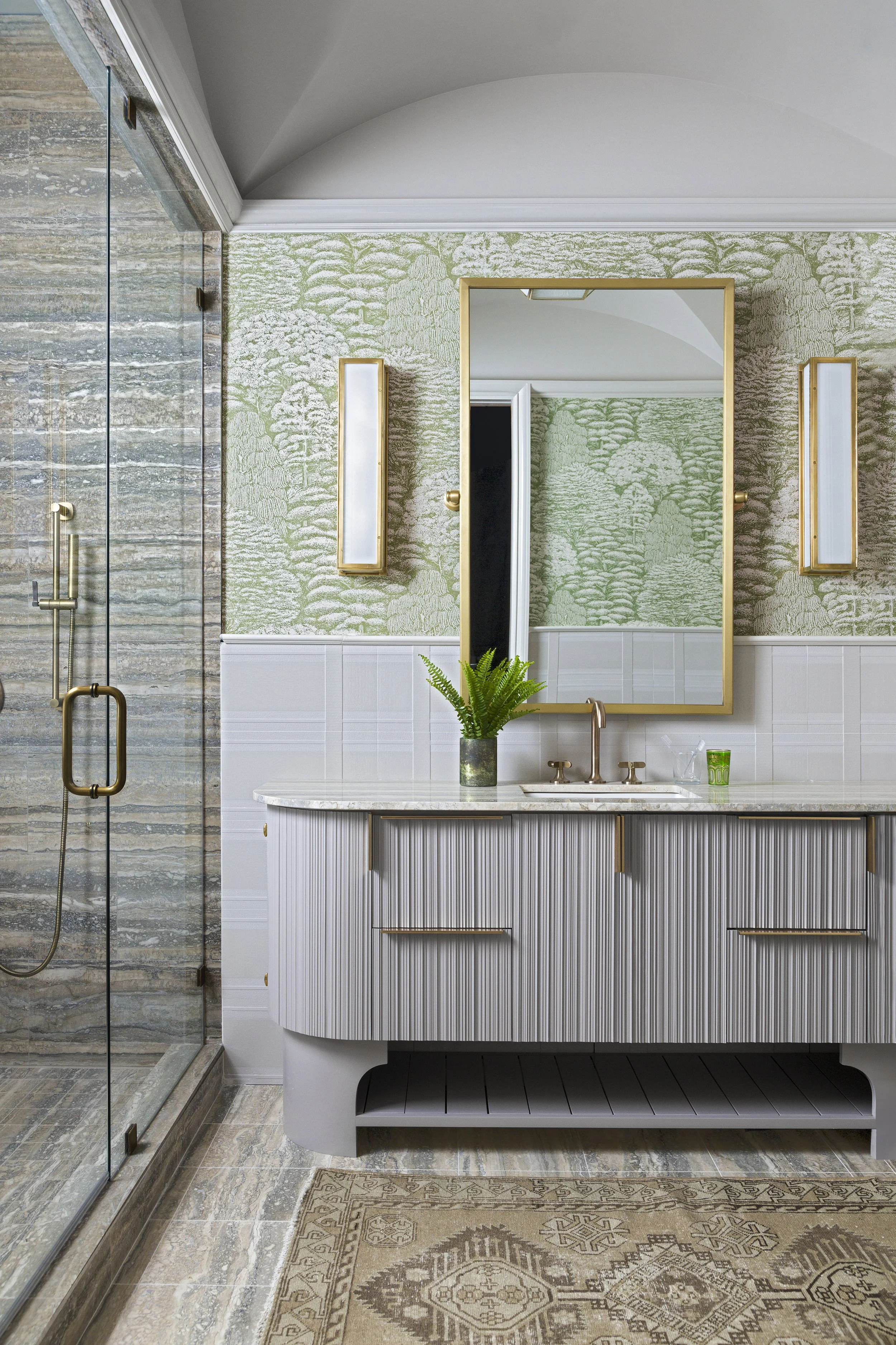 Custom bathroom vanity with fluted cabinetry, brass fixtures, botanical wallpaper, and glass-enclosed shower.