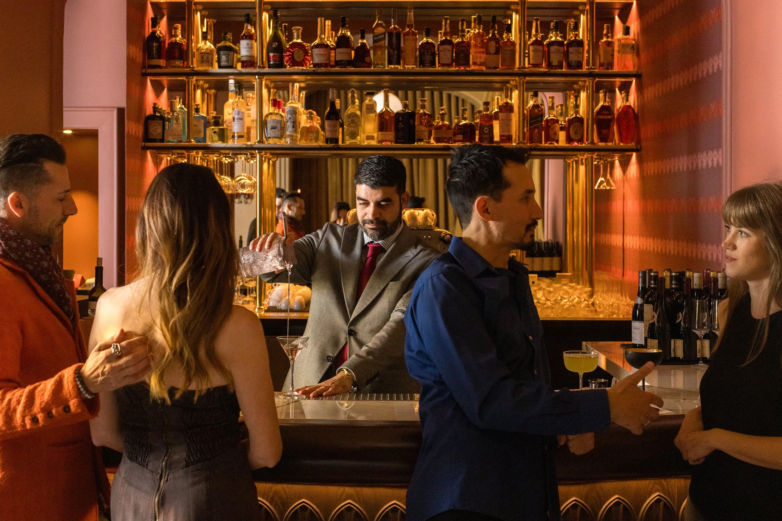 Bartender preparing a cocktail behind a warmly lit bar as guests gather in an intimate hospitality setting.
