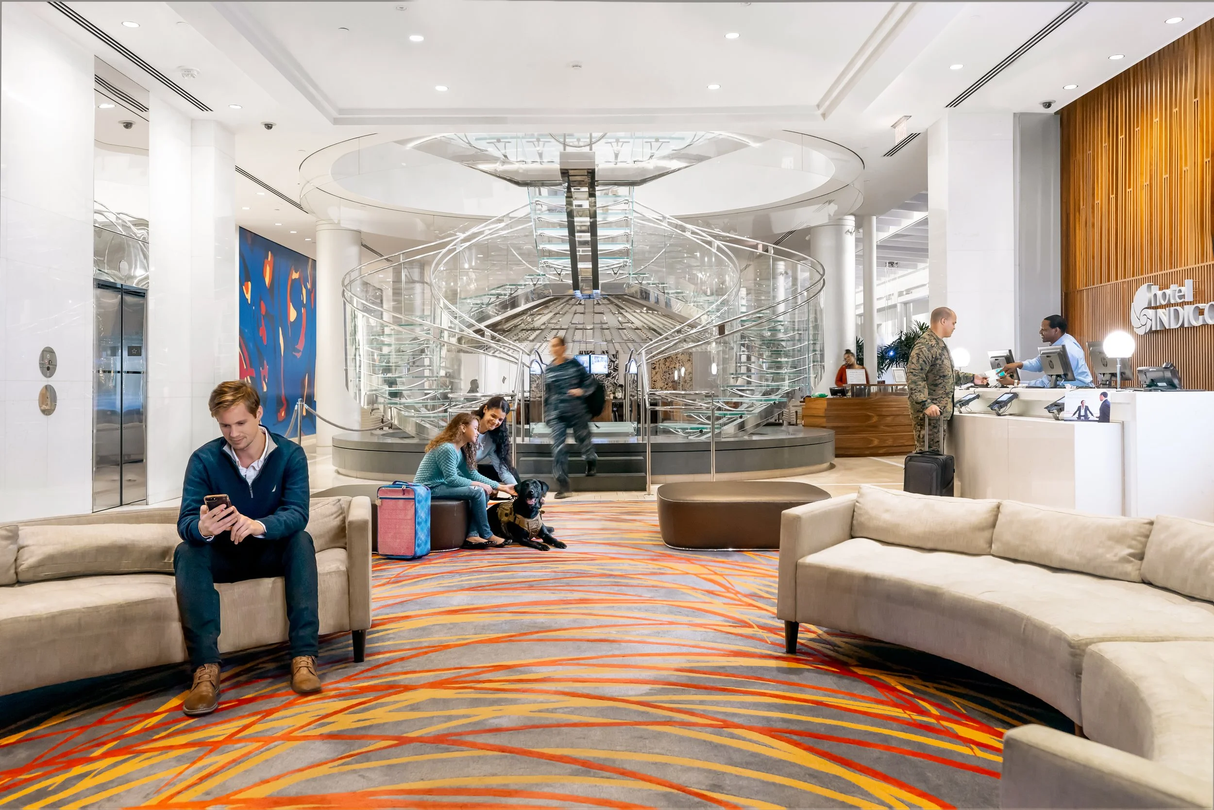Modern hotel lobby with sculptural glass staircase, curved seating, patterned carpet, and guests interacting at the reception desk.