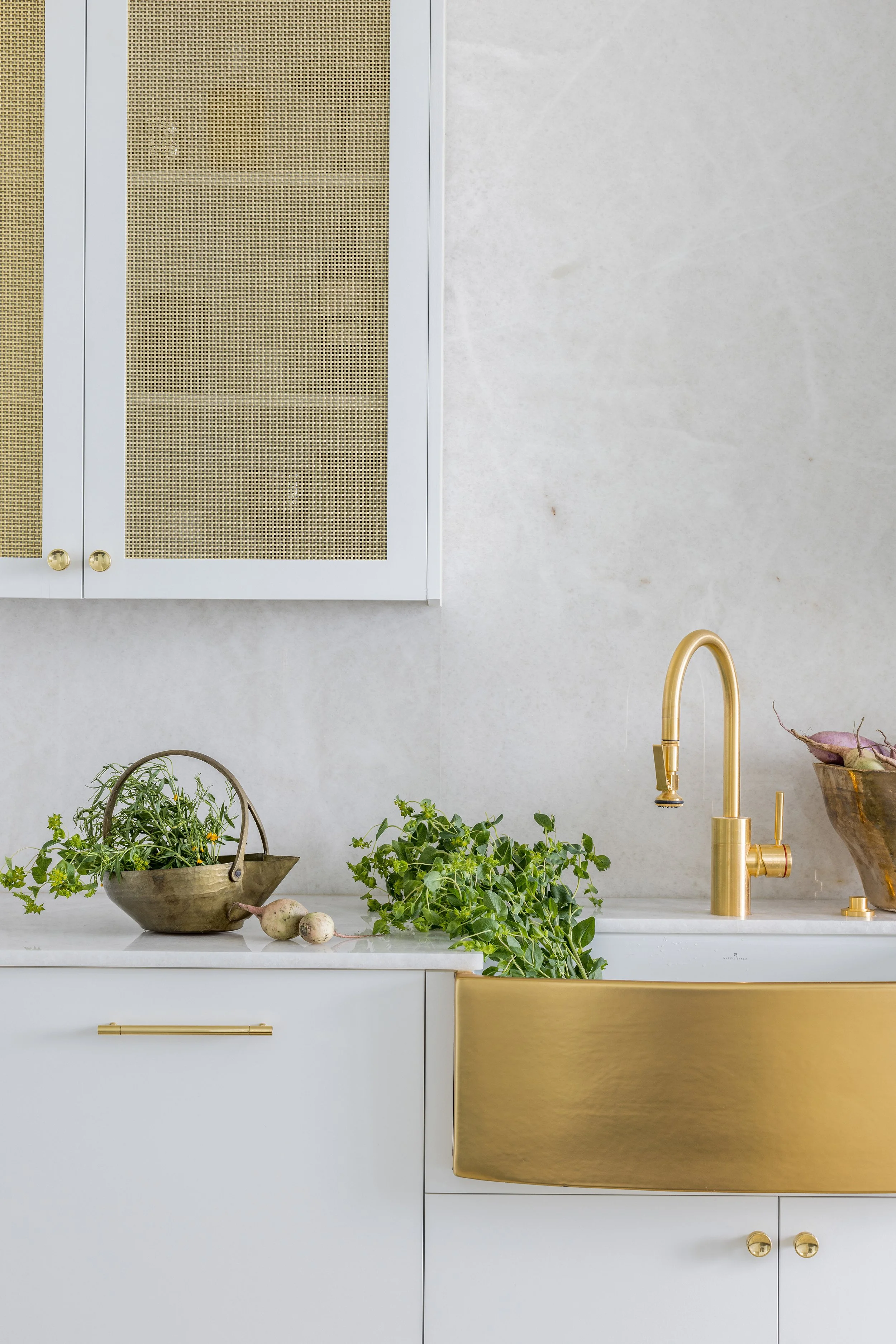 Kitchen detail with brass apron-front sink, white cabinetry, stone backsplash, and woven cabinet inserts.
