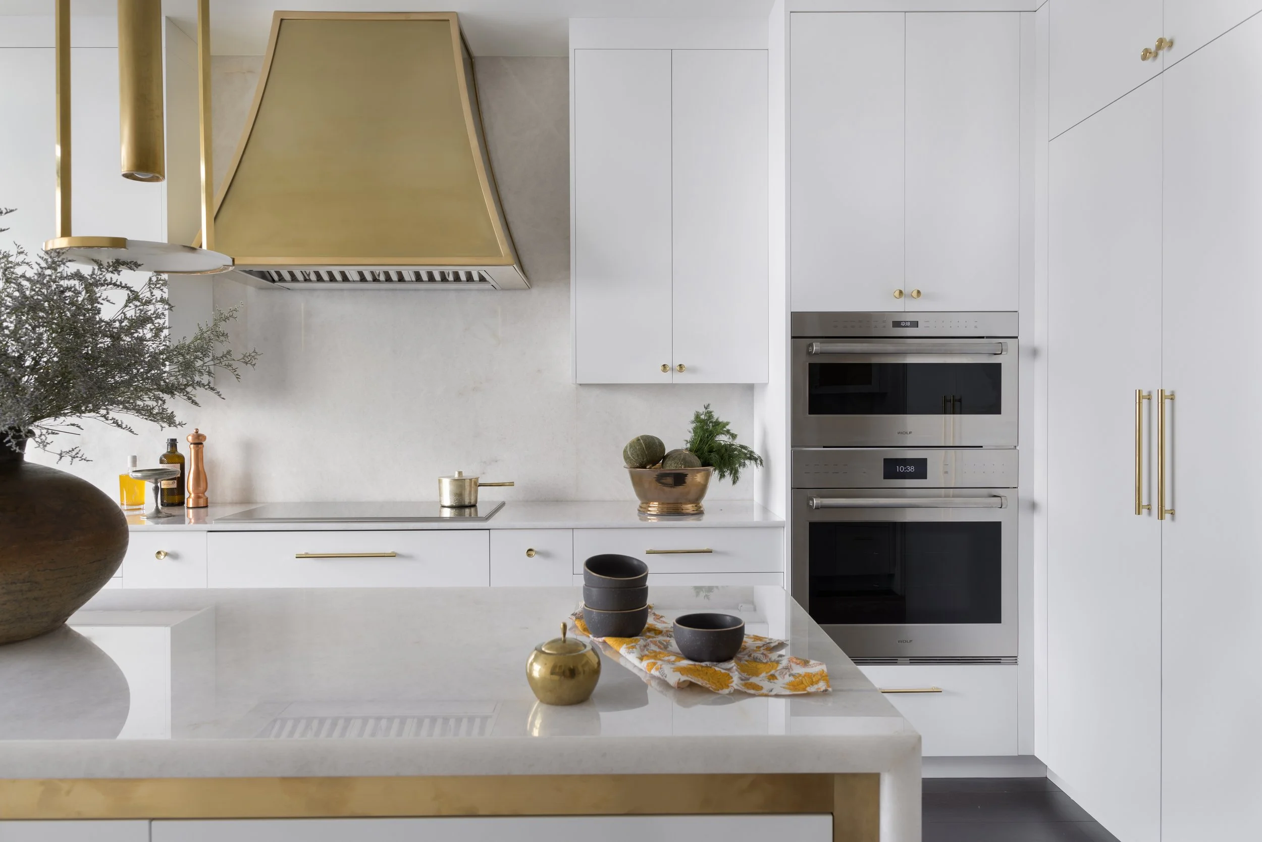 Modern kitchen with custom white cabinetry, brass range hood, integrated wall ovens, and stone countertops.