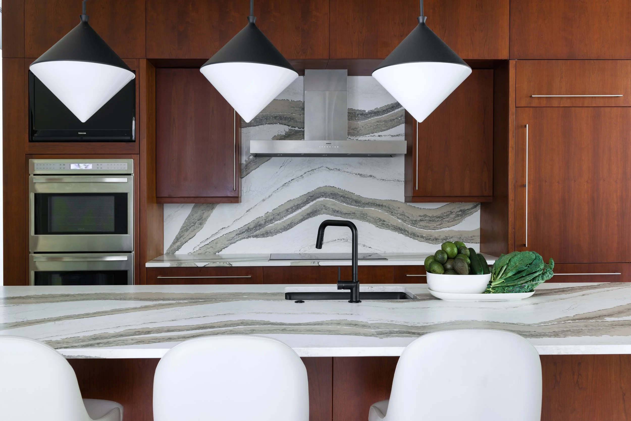 Modern kitchen with waterfall stone island, warm wood cabinetry, black faucet, and sculptural pendant lighting.
