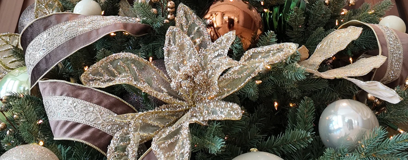 Close-up detail from a bronze and gold-adorned Christmas tree, a part of one of our holiday decorating projects. Features metallic ornaments and shimmering floral accents, velvet and lace ribbon, and traditional-style round balls.