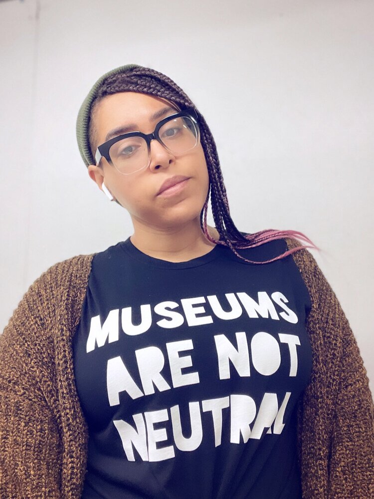 Jillian Marie Browning self portrait - Black femme person with long braids and large glasses, wearing a black shirt with white letters that read 