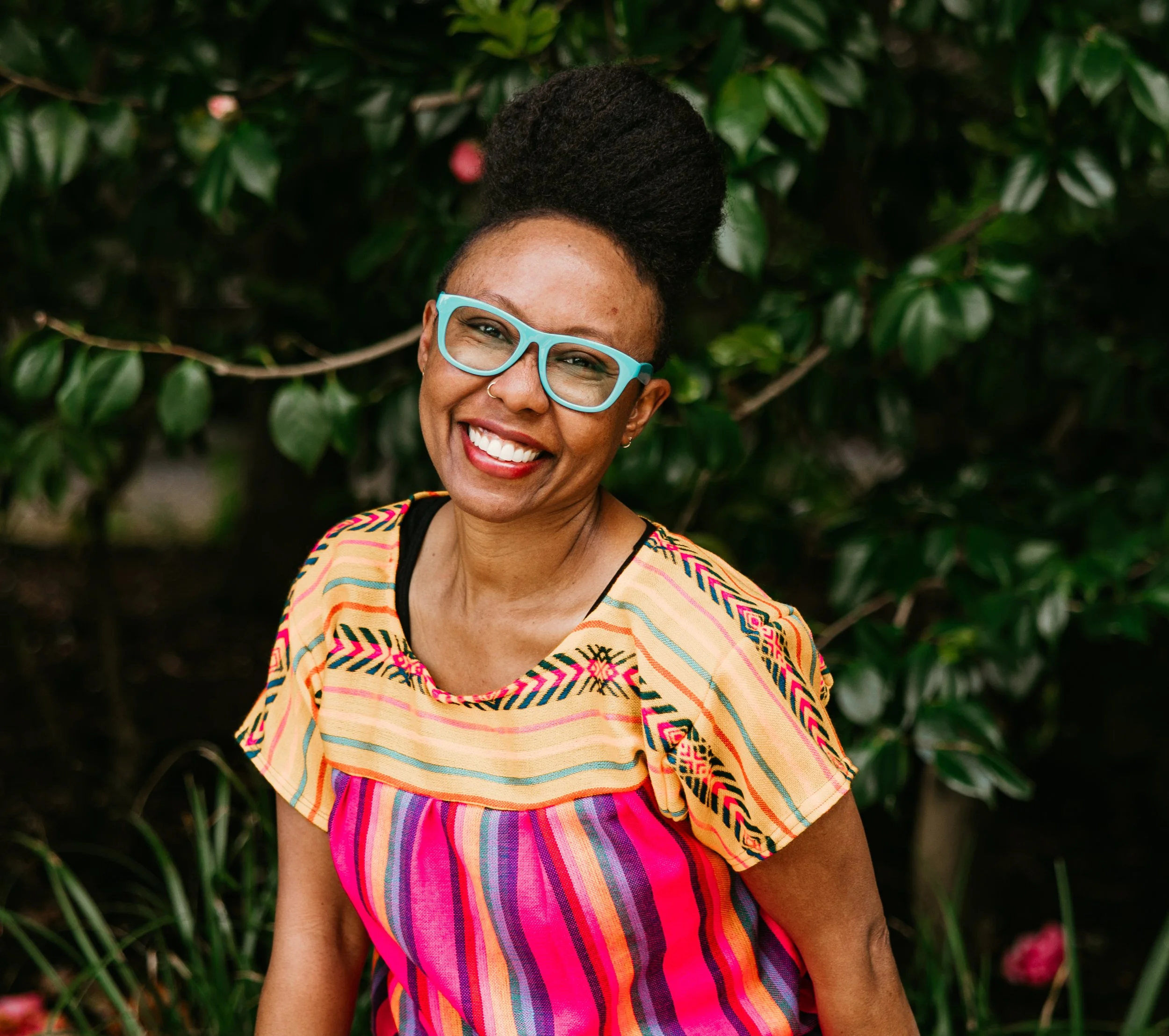 Kat, a Black woman with an afro pompadour and teal glasses smiles in front of a camellia tree, wearing a bright yellow and pink summer blouse