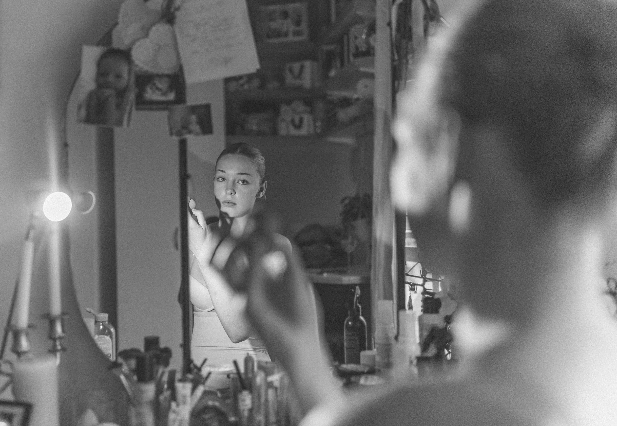 Woman applying makeup in mirror, surrounded by cosmetics and a lit lamp.