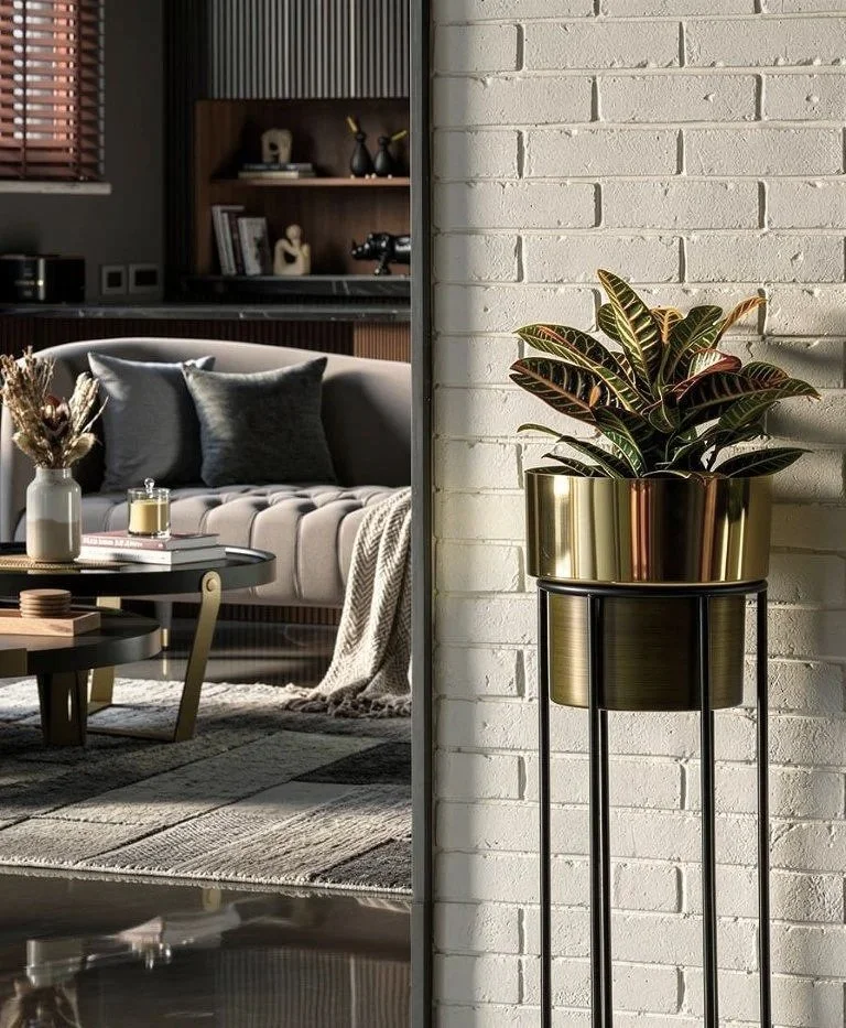 Interior living room with a beige sofa, dark pillows, a low round coffee table, a bookshelf with decor, and a large plant in a gold pot on a stand by a white brick wall.