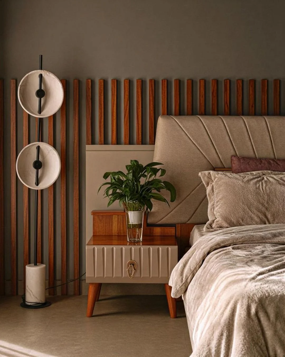 A bedside table with a potted plant, a bed with a beige headboard and pillows, wooden slat wall, and modern floor lamp with dual circular shades.