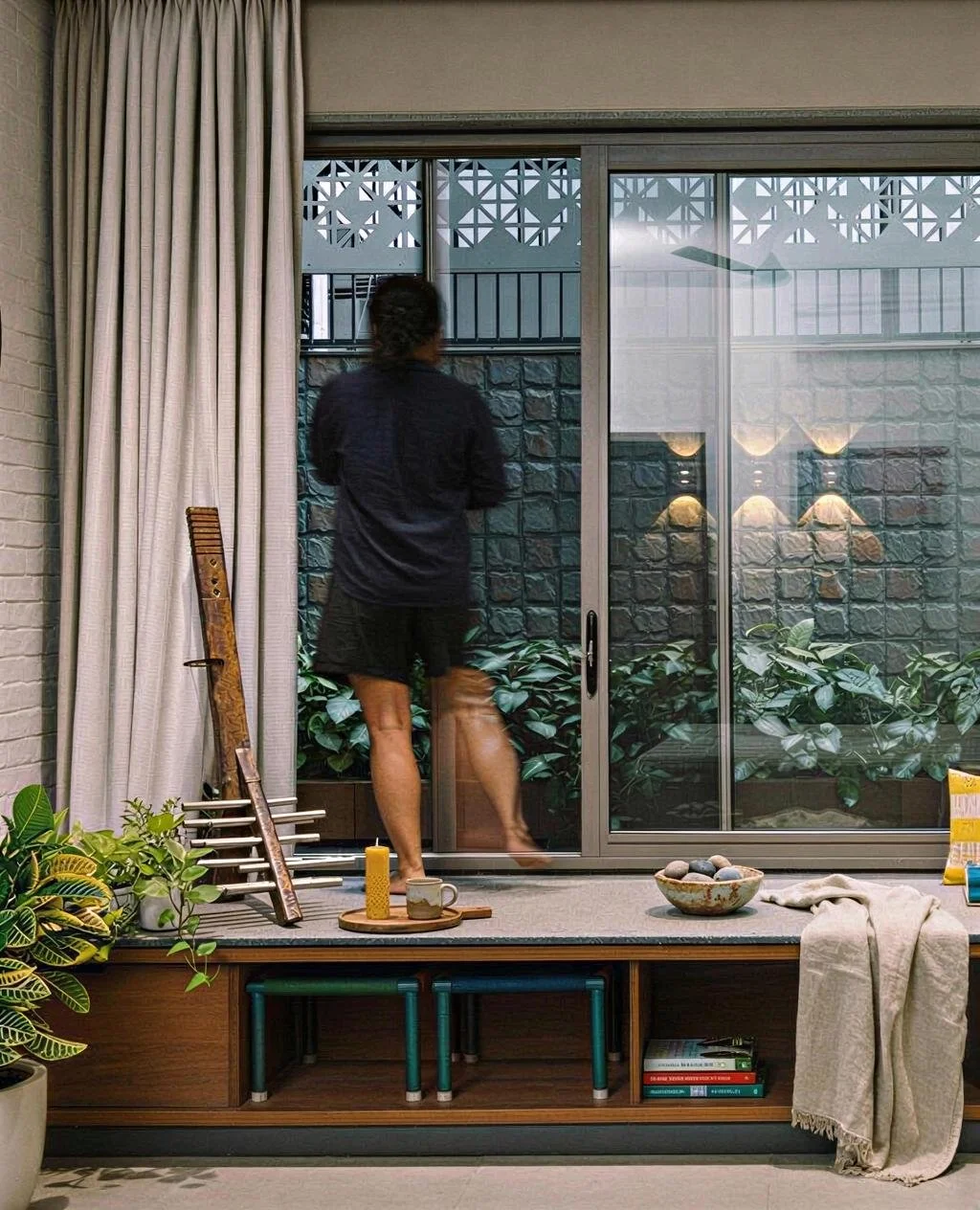 A person standing by a sliding glass door, looking outside, with various indoor plants, a bowl of stones, a candle, a cup, and books inside the room.