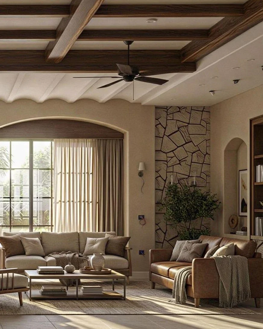 A living room with beige walls, a large arched window with curtains, a beige sofa, a brown leather sofa, and a small coffee table with books and decorative items. There is a ceiling fan, a stone accent wall, and a built-in bookcase.