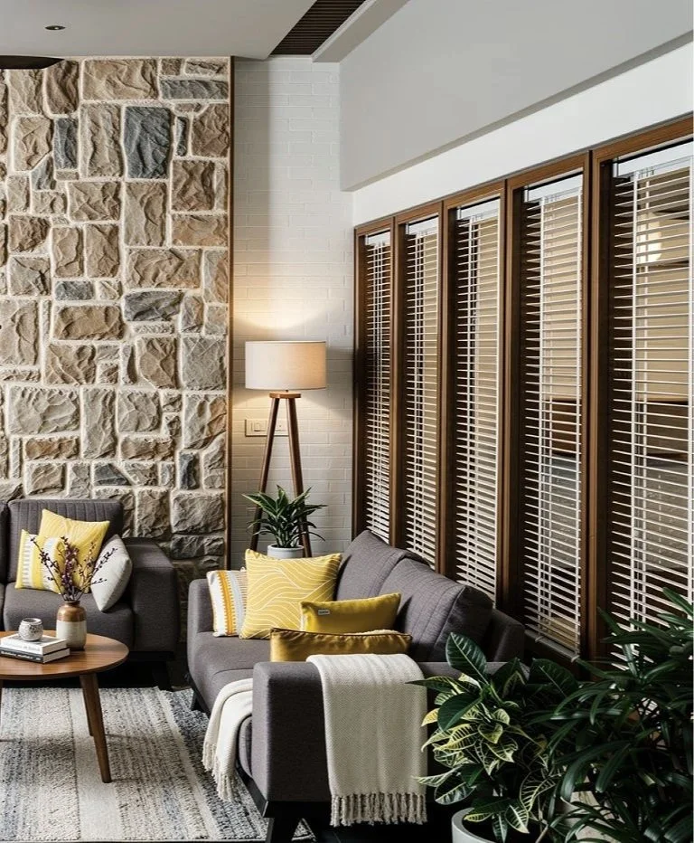 Living room with stone accent wall, large wooden window shutters, modern gray sofa with yellow throw pillows, a wooden coffee table with decorative items, a tall tripod floor lamp, a potted plant, and a patterned area rug.