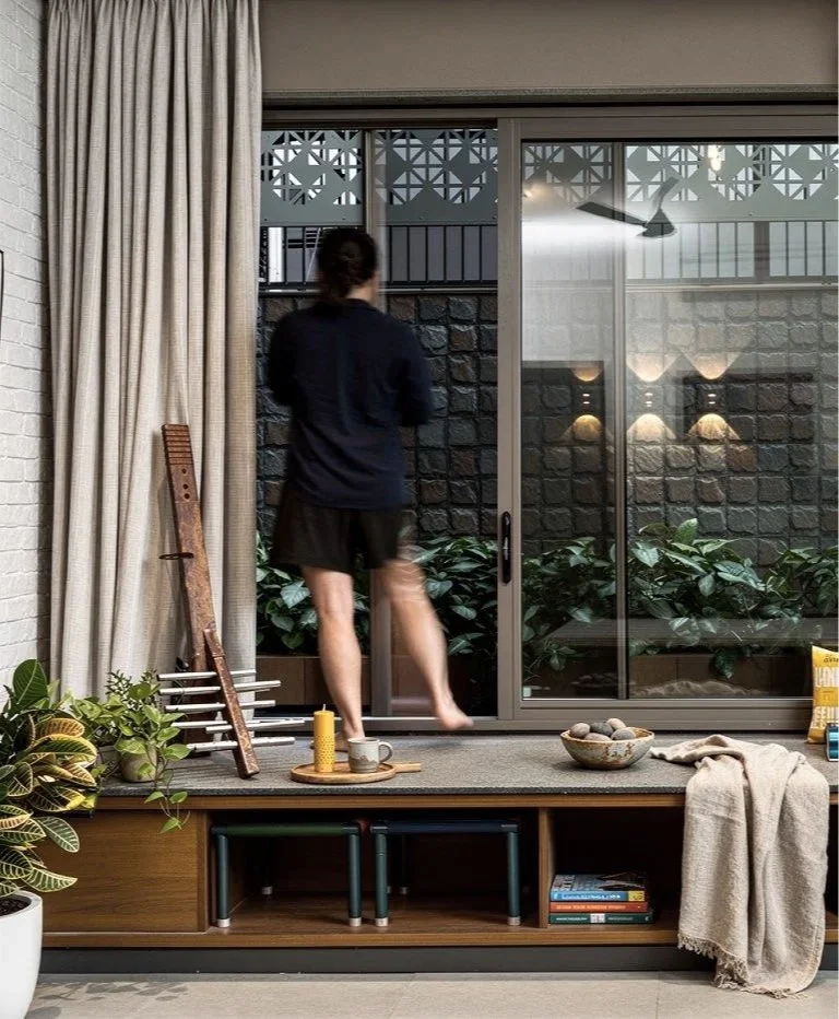 A person standing by a sliding glass door, looking outside, with various indoor plants, a bowl of stones, a candle, a cup, and books inside the room.