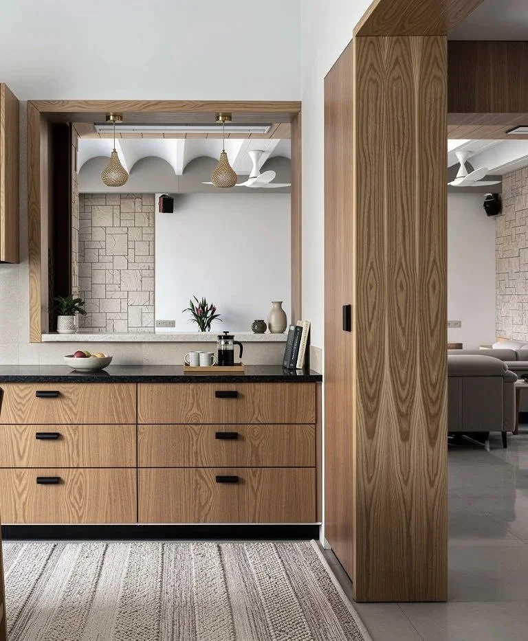 Interior view of a living space with a kitchen area featuring wooden cabinets and countertops, a decorative cutout wall with potted plants and vases, and a glimpse into a living room with a sofa and modern ceiling fans.
