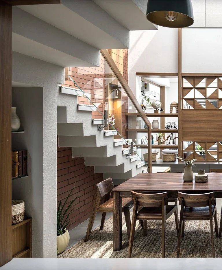A modern dining room with a wooden table and chairs, decorative vases, a staircase with a glass railing, and built-in shelving filled with decorative items and plants.