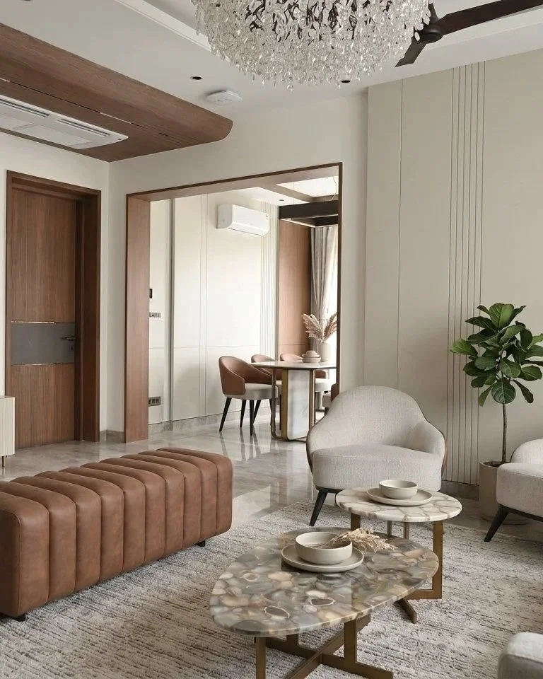 Modern living room with beige armchair, brown tufted ottoman, marble-topped side tables with bowls, large potted plant, and a chandelier, with open view into a dining area with a round table, four chairs, and window curtains.