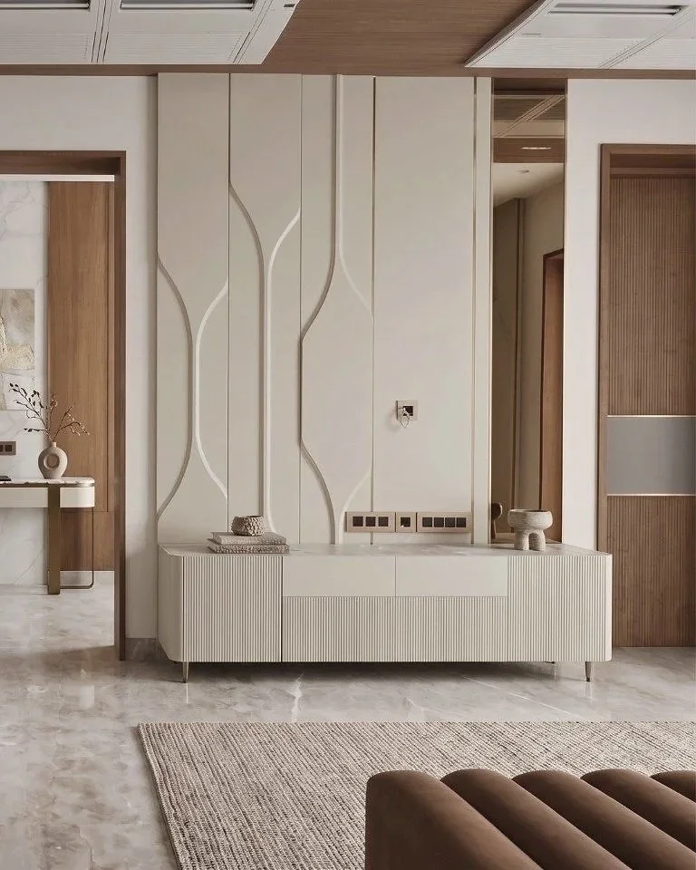 Modern living room with beige textured walls, a white textured TV console, and decorative wall panels. There is a portion of a brown sofa in the foreground, a beige rug on a marble floor, and some decorative objects including a small sculpture and a 