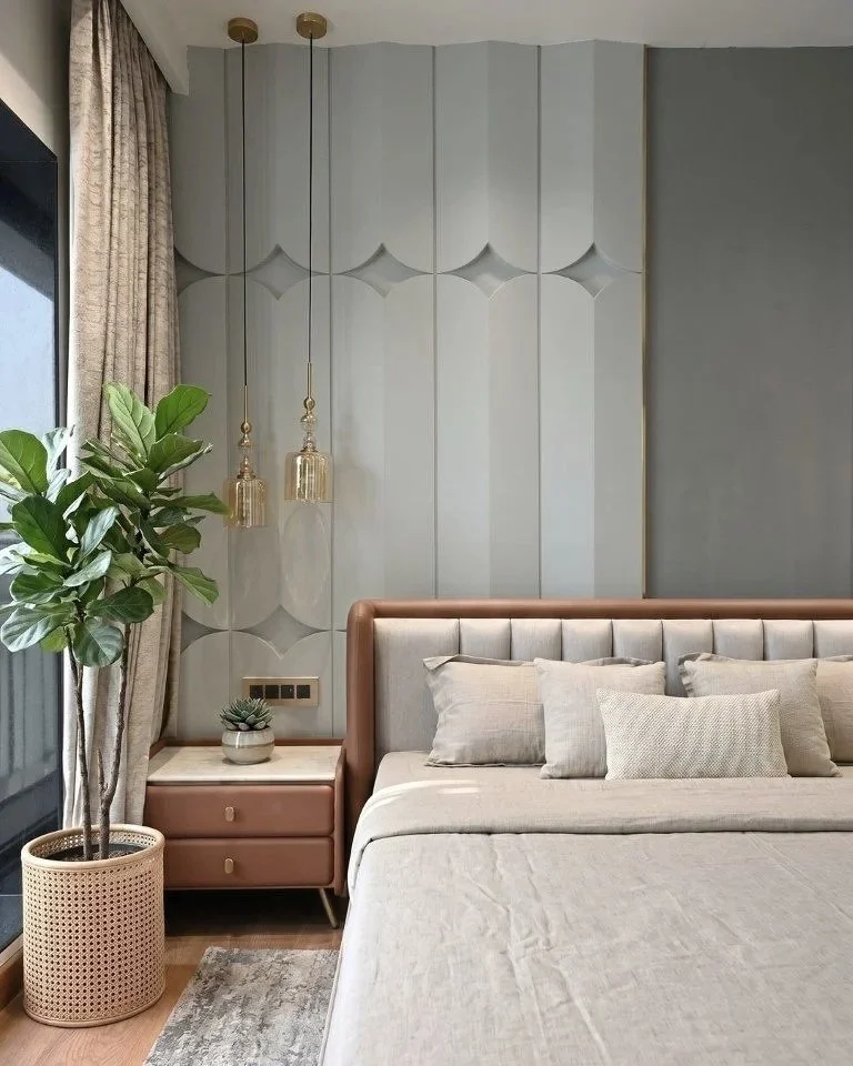 Modern bedroom with light gray wall panels, beige bedding, and multiple pillows. A large leafy plant in a woven basket is near a window with beige curtains. A pink nightstand with a small plant and gold hanging pendant lights is beside the bed.