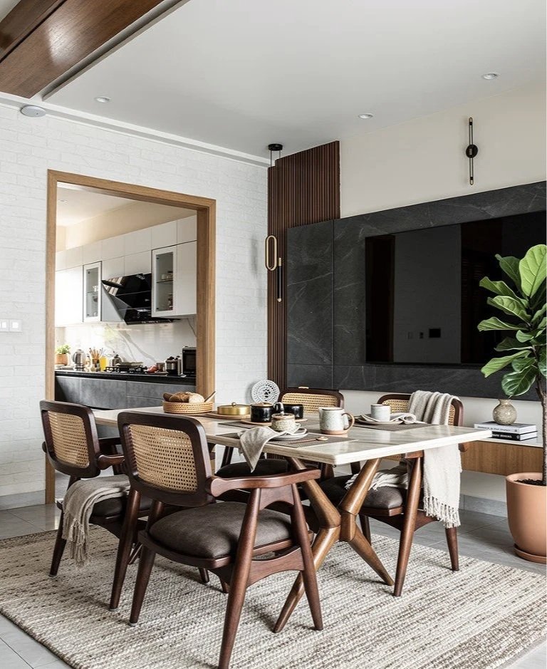 A modern dining room with a wooden table set for breakfast and six chairs, adjacent to a kitchen with white cabinets and black range hood, decorated with indoor plants and contemporary decor.