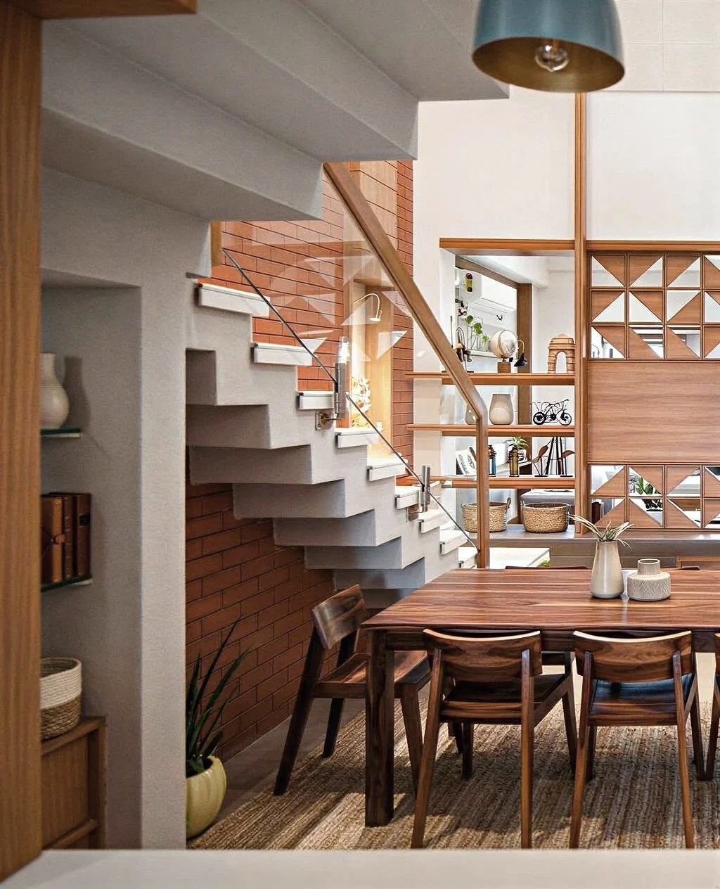 Interior view of a modern dining area with a wooden table, six matching chairs, and decorative vases on the table. A staircase with concrete steps and a glass railing is visible, along with wooden and brick wall accents, open shelves with decor, and 