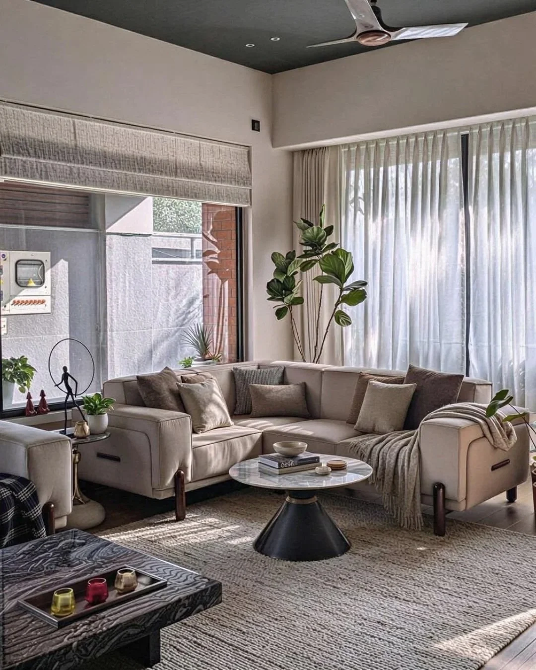 Bright living room with large cream sectional sofa, various pillows, and a round marble coffee table. Large windows with sheer curtains and tall indoor plants add to the cozy atmosphere. Decor includes small sculptures, books, and candles.